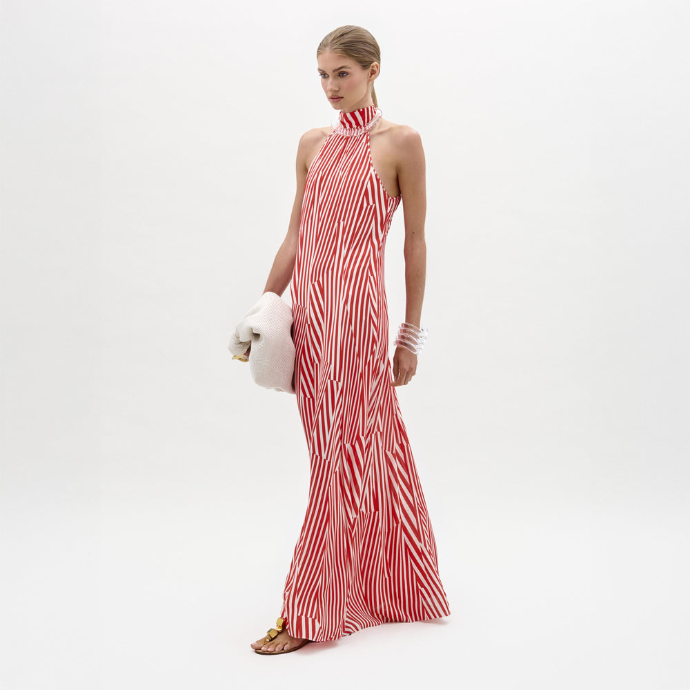 A woman wears the Koa Dress, a red and white striped halter neck maxi with an A-line silhouette, standing against a plain white background while holding a white towel and wearing sandals.