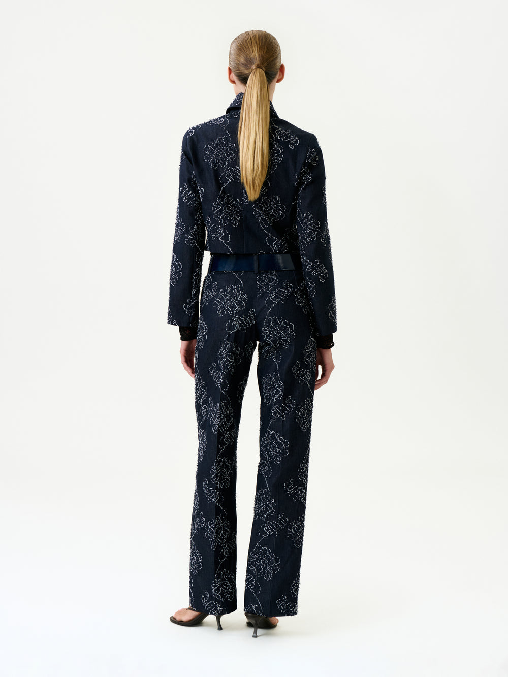 A woman with a low ponytail stands facing away, wearing the Ulla Pant—a dark blue pantsuit with a white floral pattern and black heels—made in Colombia, against a plain white background.