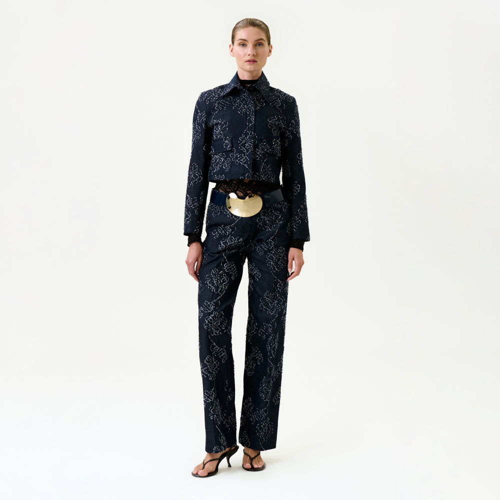 A woman poses against a plain white background, wearing the Ulla Pant and matching dark patterned jacket made from luxurious Italian fabric, paired with black heeled sandals and a large gold statement belt.