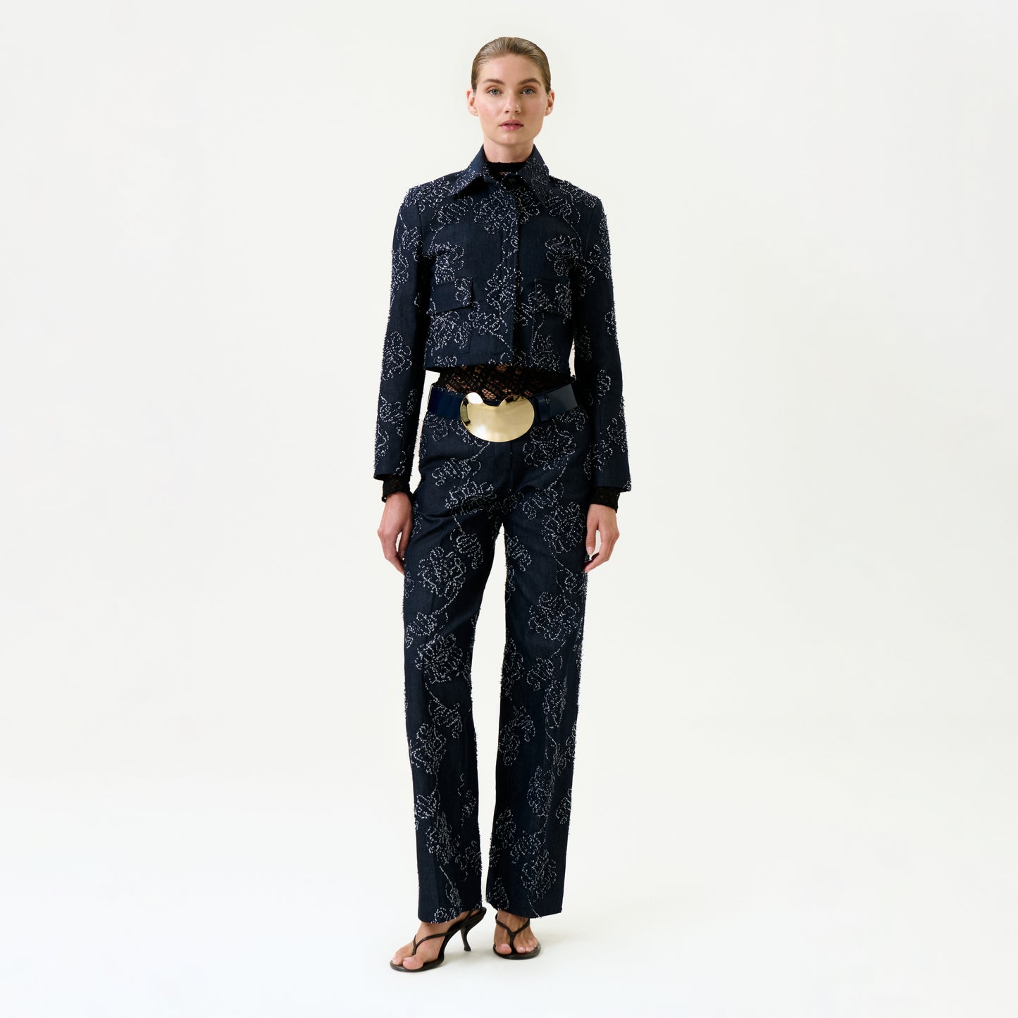 A woman poses against a plain white background, wearing the Ulla Pant and matching dark patterned jacket made from luxurious Italian fabric, paired with black heeled sandals and a large gold statement belt.