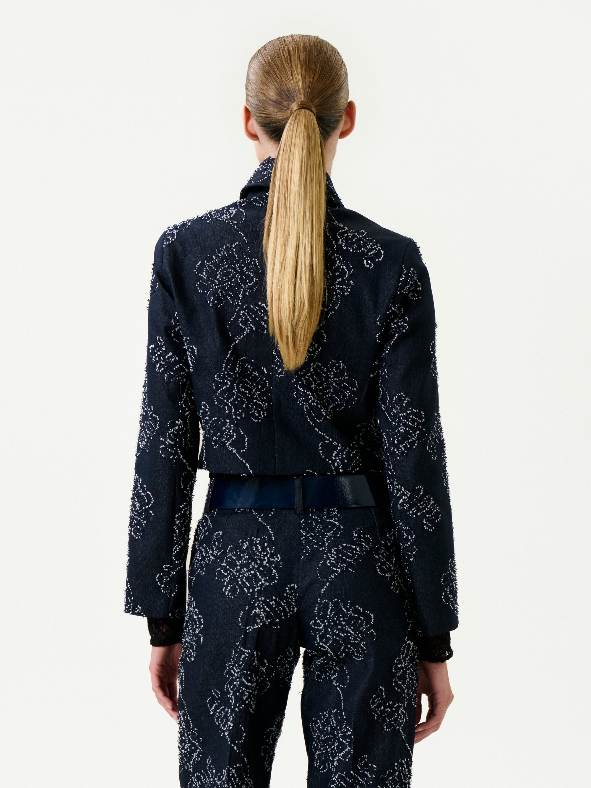 A person with long, straight blonde hair in a low ponytail is seen from behind wearing the Zulma Jacket—a dark, fitted Italian fabric jacket with a subtle light floral pattern and true-to-size fit. The background is plain white.
