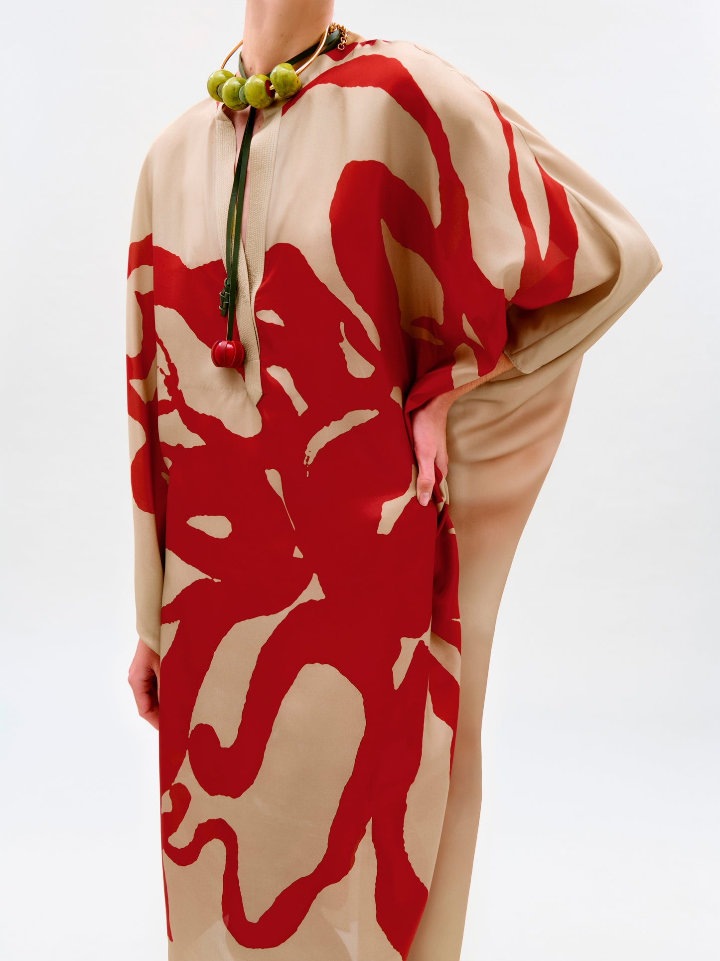 Wearing the Elea Tunic—a loose, beige maxi dress crafted from 100% Italian silk with bold red abstracts—paired with a chunky green necklace, they pose confidently against a plain backdrop.
