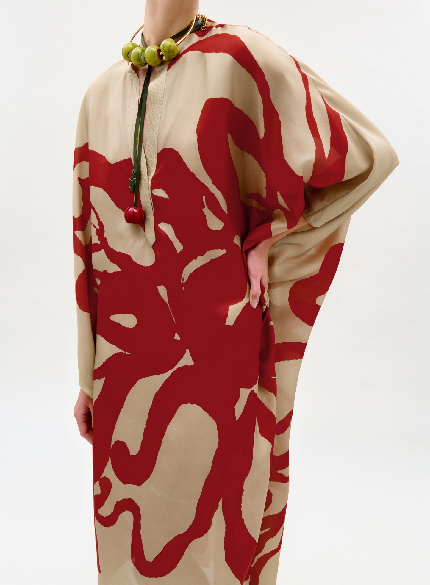 A person models the Elea Tunic, a loose beige maxi dress with bold red abstract Italian patterns, paired with a chunky green bead necklace featuring a cherry pendant, standing with one hand on their hip against a white background.