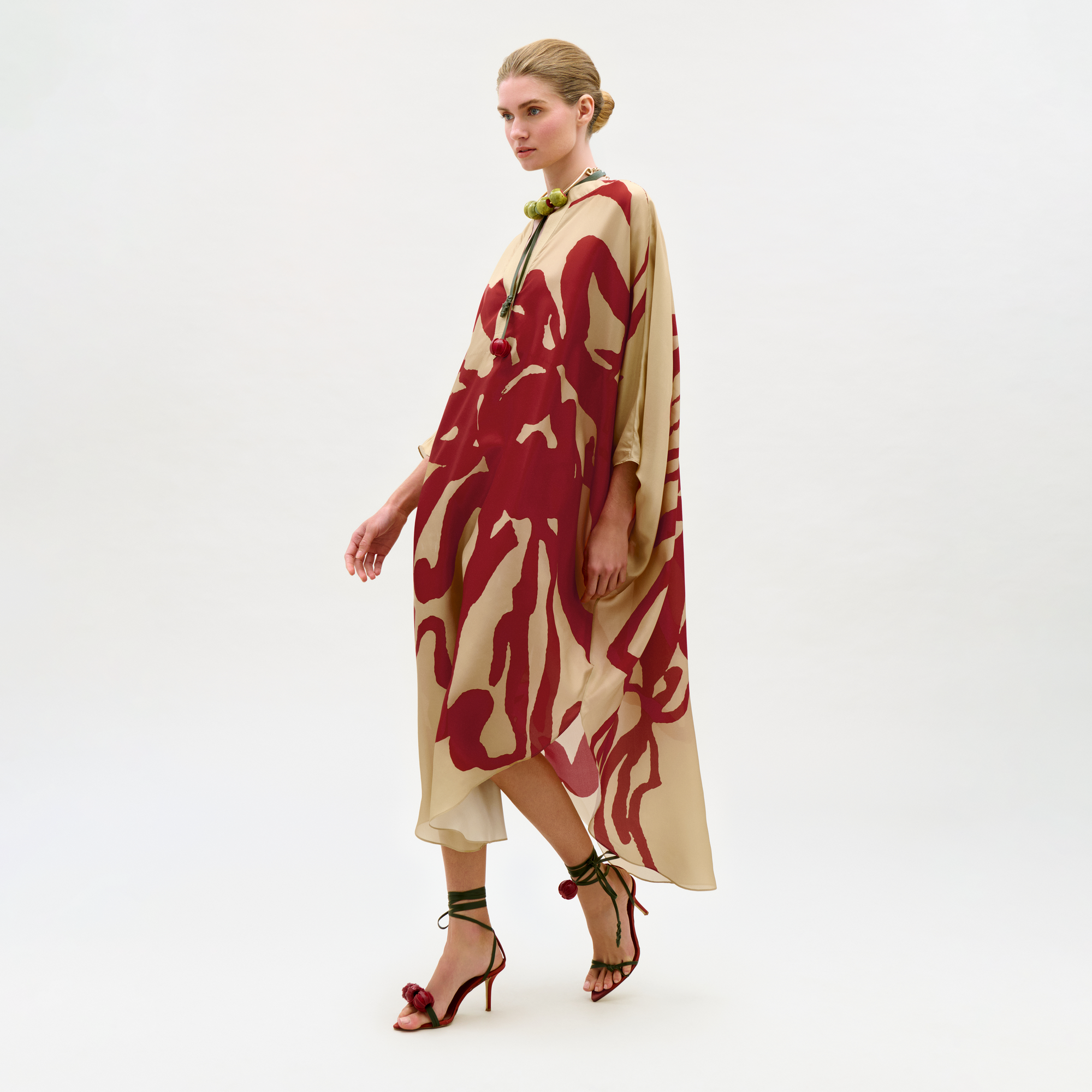A woman models the Elea Tunic, a loose, cream maxi dress with bold red abstract patterns made from Italian fabric, styled with a green necklace and red high-heeled sandals against a plain white background.