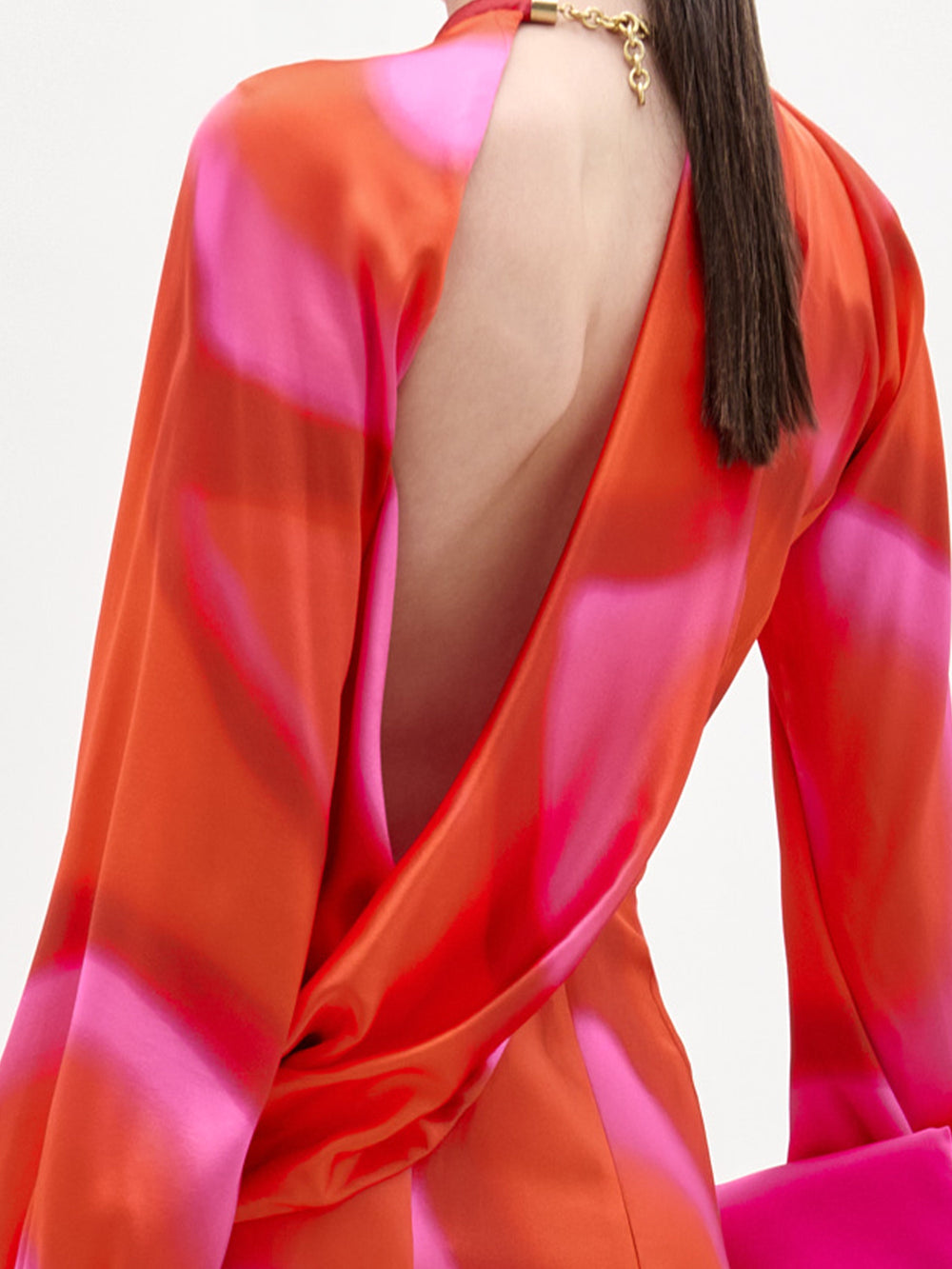 A woman stands against a plain background wearing the Muna Dress, a long-sleeve red and pink patterned silk midi dress, paired with a bright pink clutch and brown sandals.