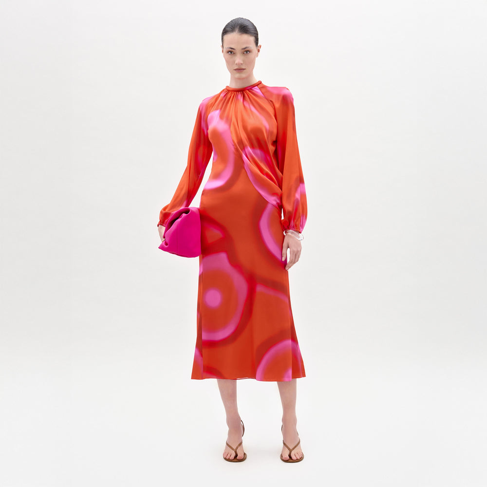A woman stands against a plain background wearing the Muna Dress, a long-sleeve red and pink patterned silk midi dress, paired with a bright pink clutch and brown sandals.