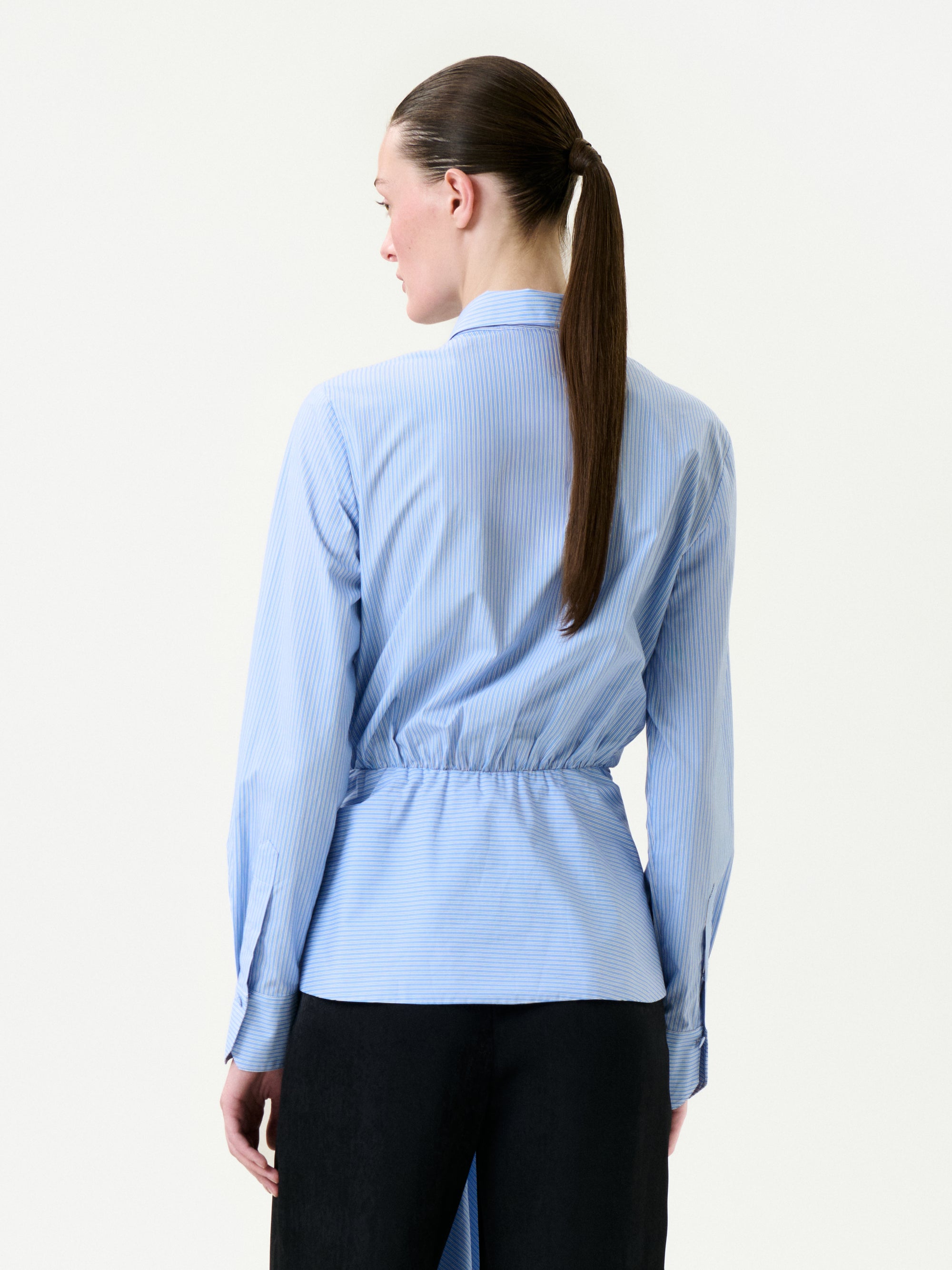 A person with brown hair in a sleek ponytail faces away, wearing the light blue, pinstriped Malen Shirt crafted from Portuguese fabric and black pants against a plain white background.