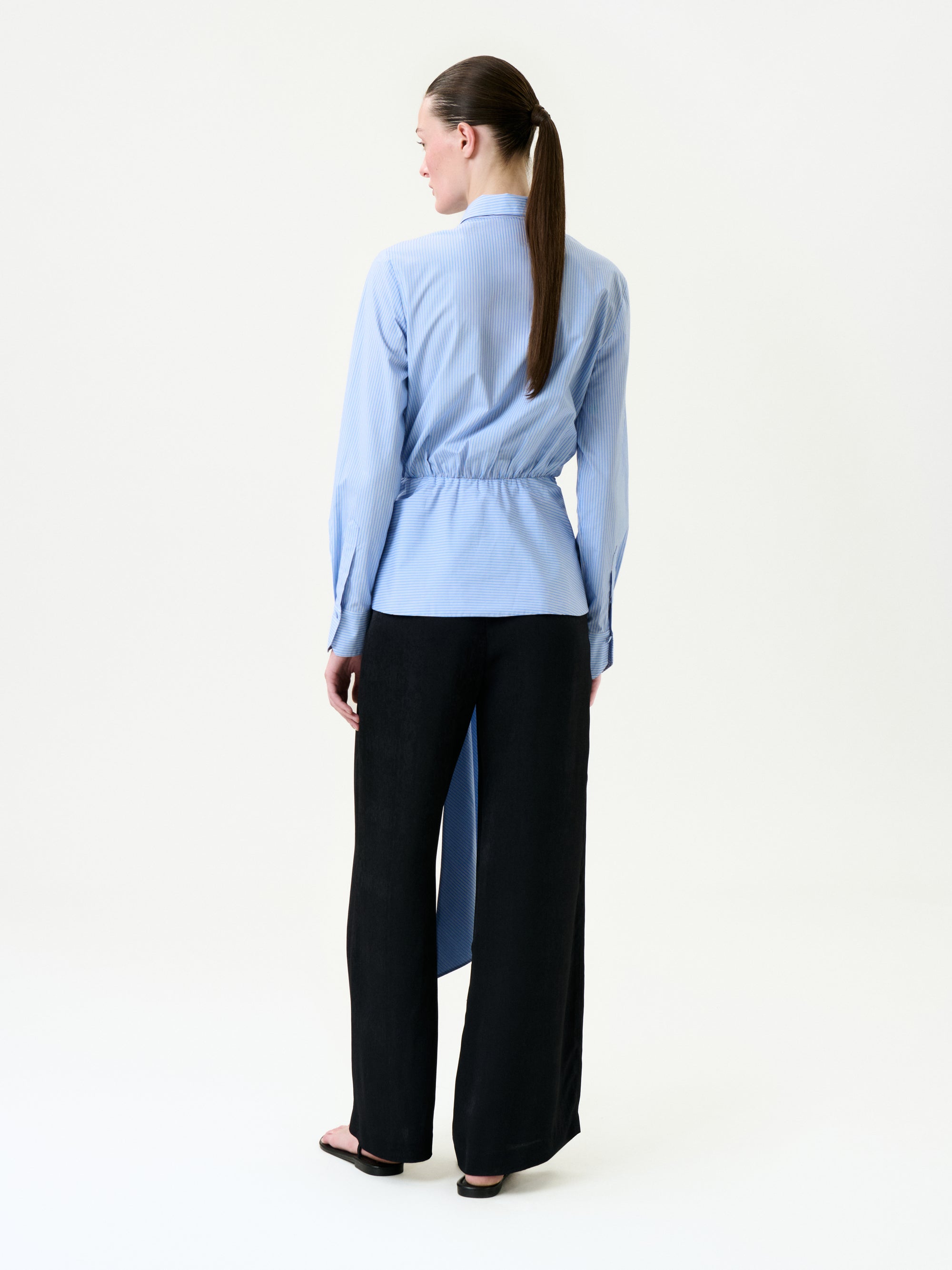 A woman with her brown hair in a low ponytail faces away, wearing loose black Ulla Pant trousers, a light blue long-sleeve 100% viscose shirt with cinched waist and asymmetrical hem, and black sandals.