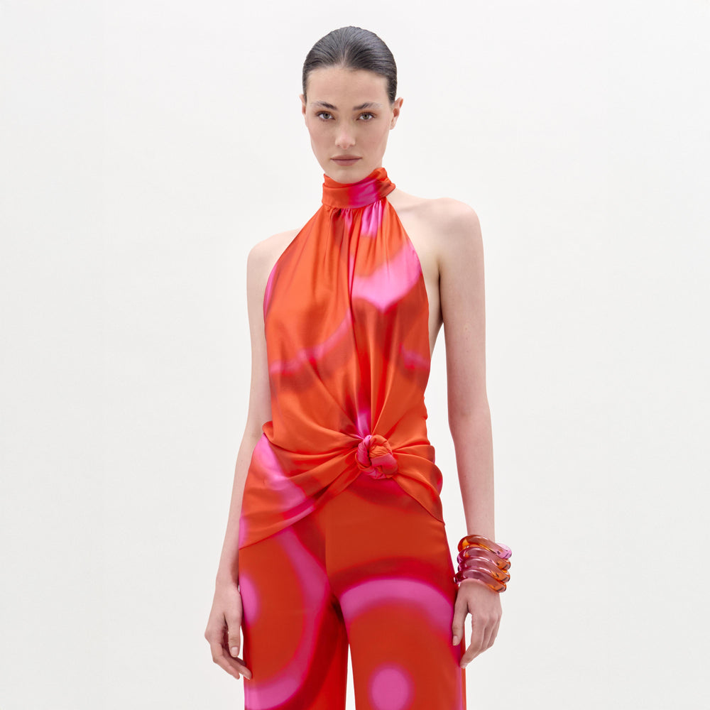 A woman wears the sleeveless, high-neck Dita Blouse featuring a red and pink pattern and knotted front, paired with matching pants and chunky bracelets, standing against a white background.