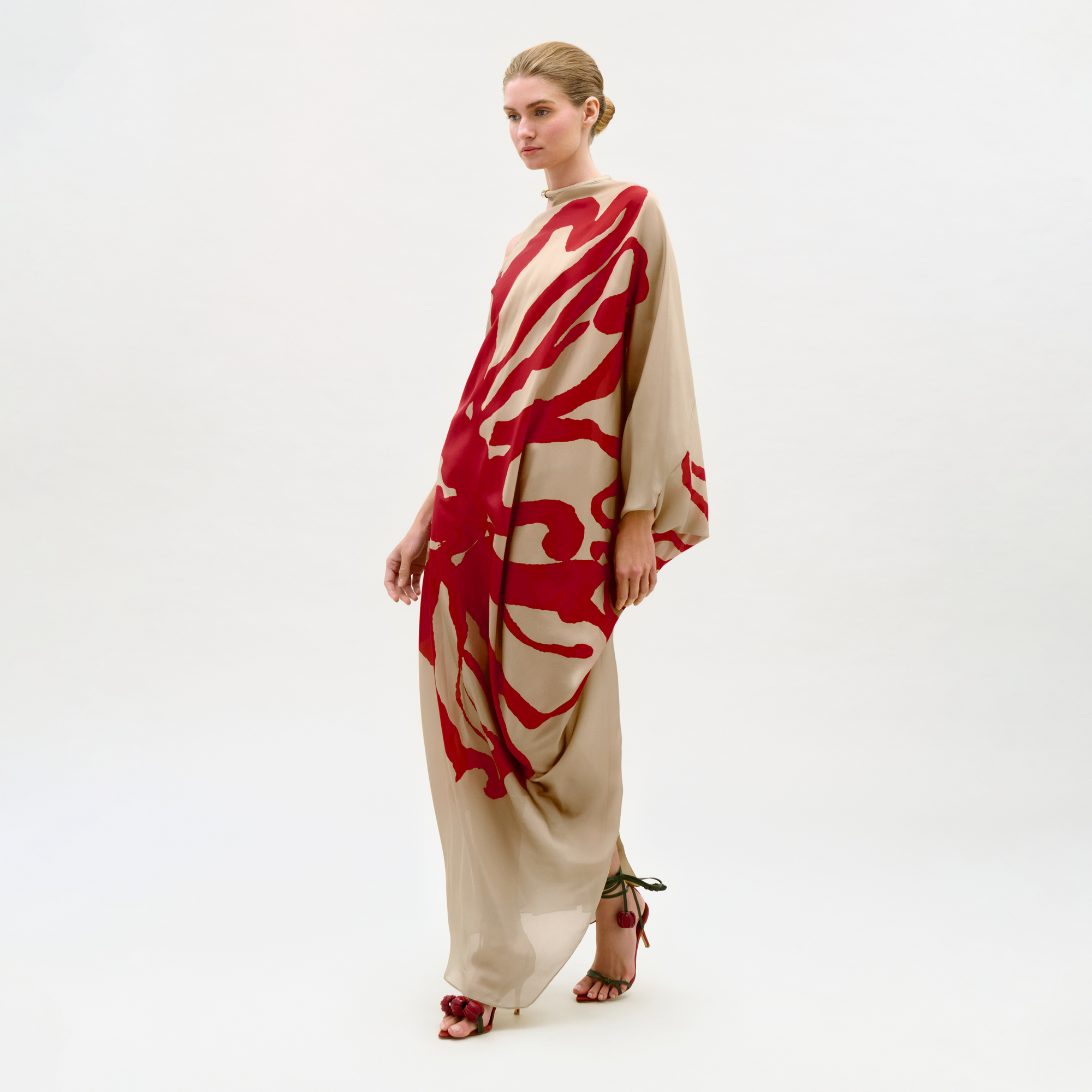 A woman models the Erica Dress, a long and loose silk maxi featuring bold red abstract patterns and billowy sleeves, styled with strappy high-heeled sandals against a plain white background.