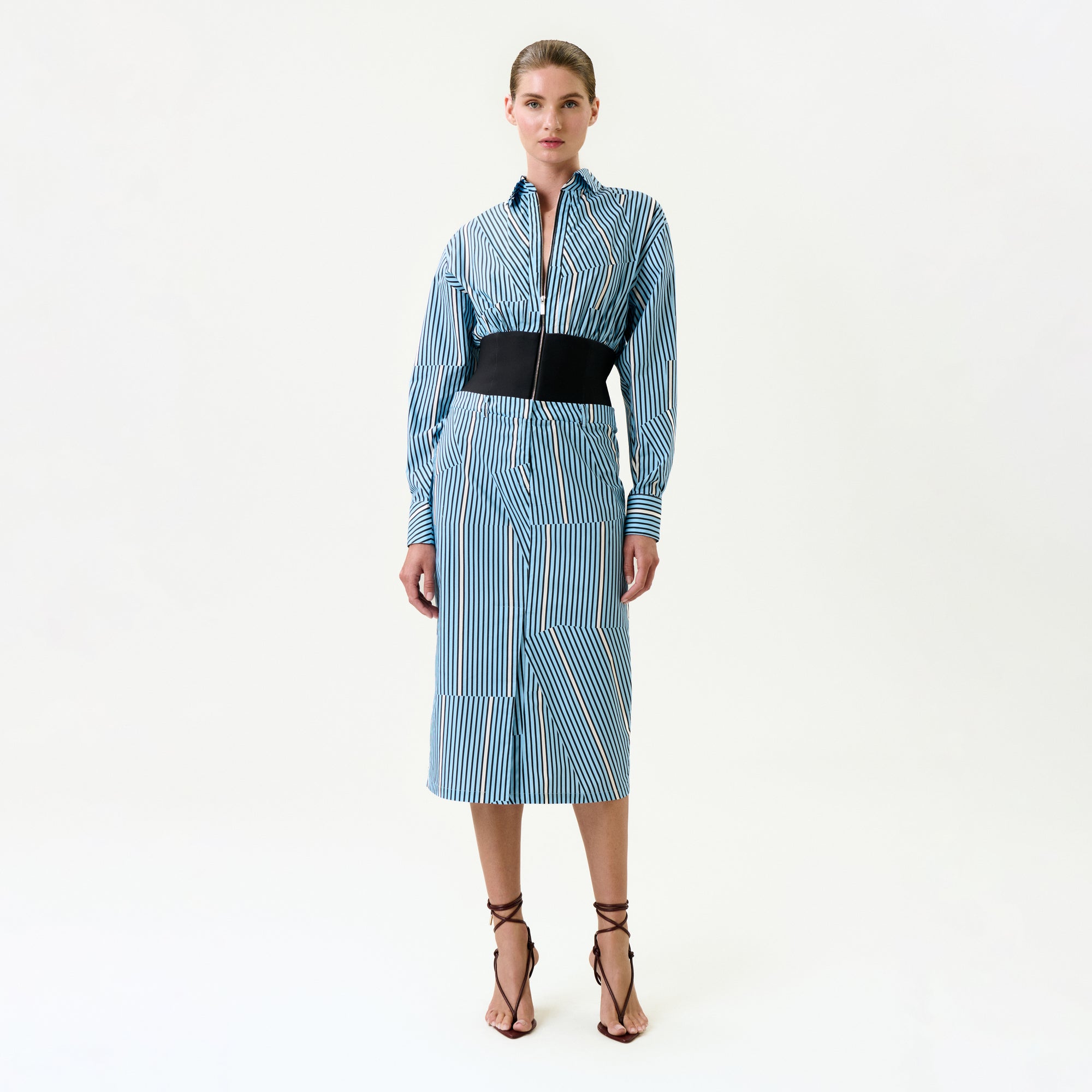 A woman poses against a white background in the Lorena Dress, a blue and white vertical striped midi made from 100% Italian cotton, with long sleeves, a black wide belt, and strappy brown high-heeled sandals.