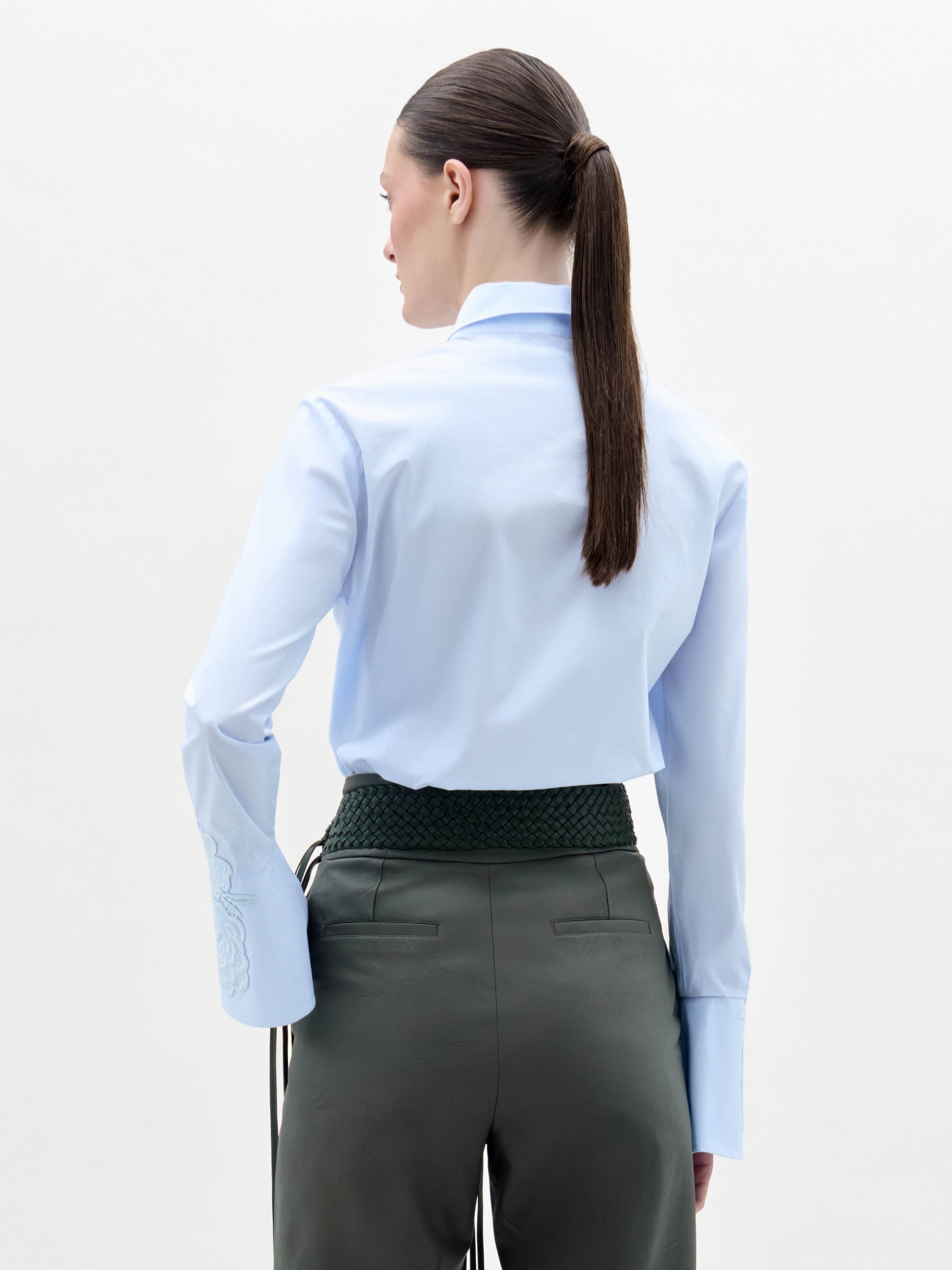 A woman stands against a plain background, wearing the Neve Blouse—a light blue collared shirt with embroidered sleeves—paired with green high-waisted pants featuring a large gold circular belt and long tassels.