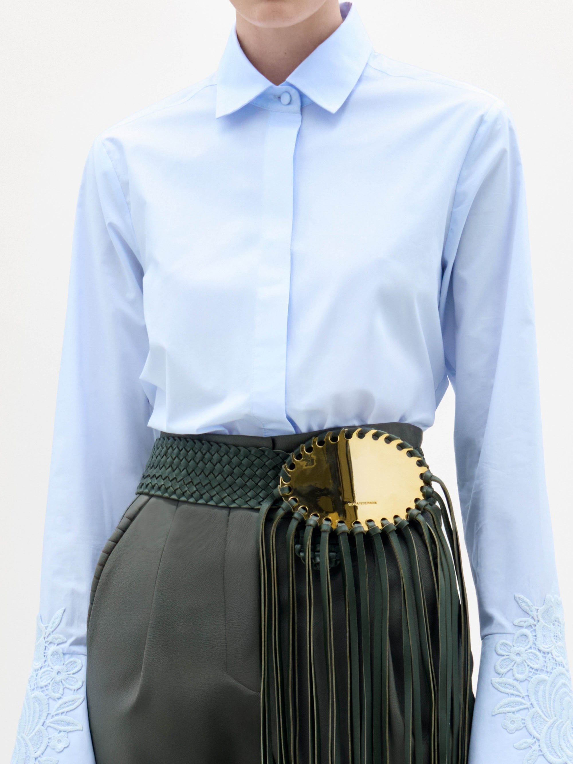 A woman stands against a plain background, wearing the Neve Blouse—a light blue collared shirt with embroidered sleeves—paired with green high-waisted pants featuring a large gold circular belt and long tassels.