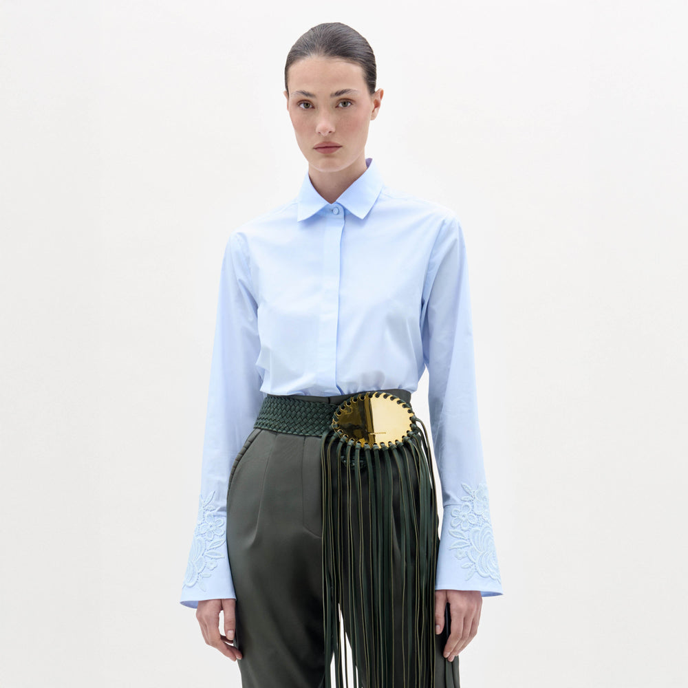 A woman stands against a plain background, wearing the Neve Blouse—a light blue collared shirt with embroidered sleeves—paired with green high-waisted pants featuring a large gold circular belt and long tassels.
