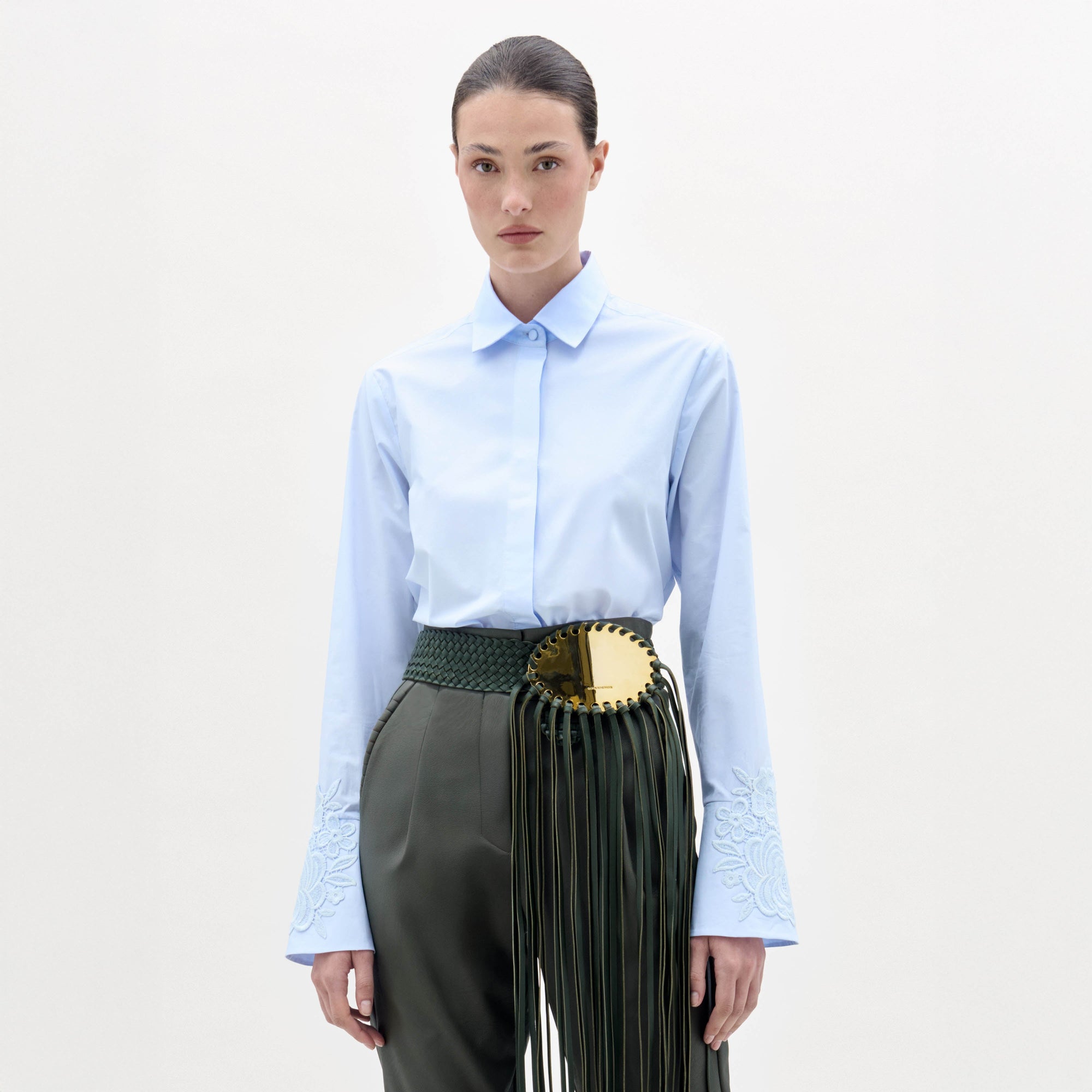 A woman stands against a plain background, wearing the Neve Blouse—a light blue collared shirt with embroidered sleeves—paired with green high-waisted pants featuring a large gold circular belt and long tassels.