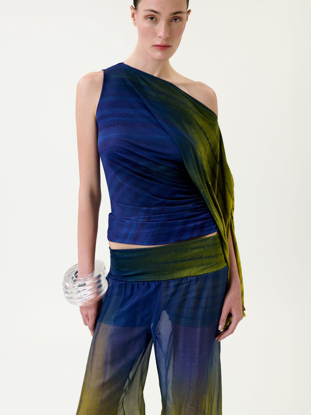 A woman wearing the Rhona Blouse—a one-shoulder blue and green top—paired with sheer matching pants and a large clear bracelet, standing against a plain light background.