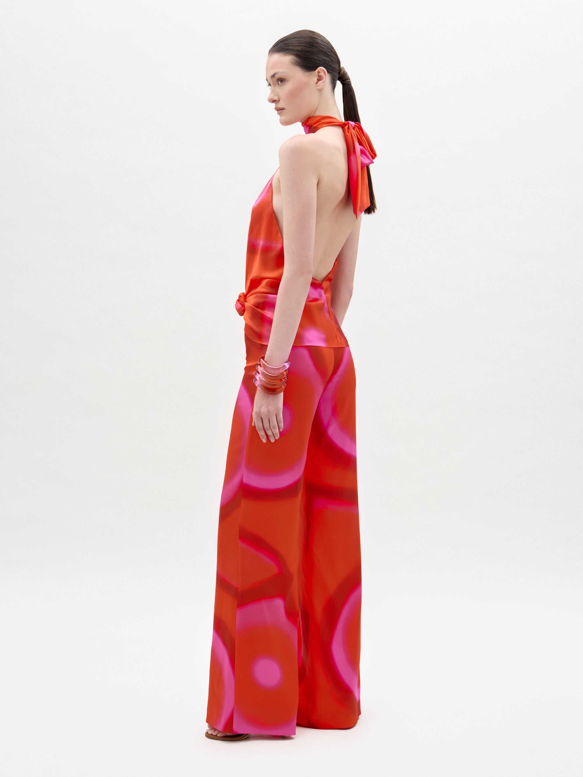 A woman stands sideways against a white background, looking over her shoulder while wearing the Andie Pant—a red and pink halter-neck backless jumpsuit with high-waisted wide-leg pants. Her straight brown hair is tied back and she accessorizes with red bangles.