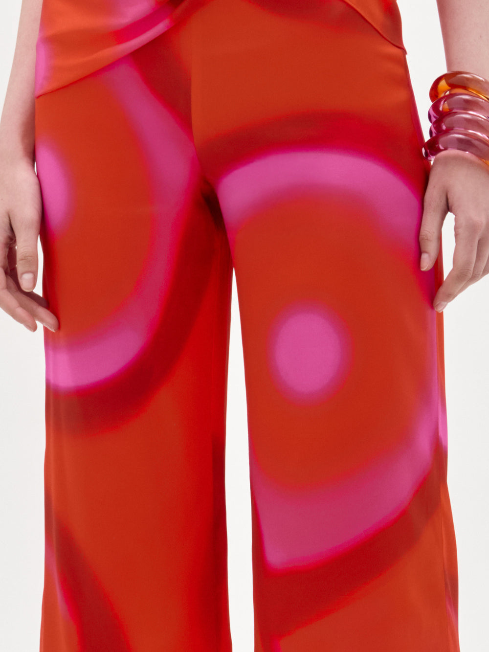A woman poses against a white background in the Andie Pant, a high-waisted, wide-leg jumpsuit with a bold red and pink circular pattern, styled with matching bracelets and sandals.