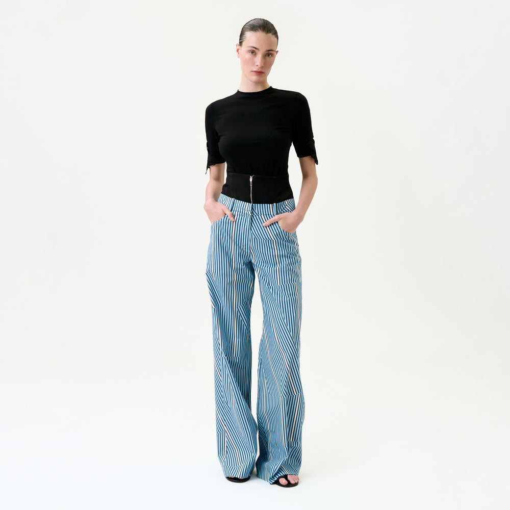 A woman stands against a white background wearing the Ruelle Pant—high-waisted, wide-leg blue and white striped 100% cotton pants—with her hands in her pockets and a fitted black top.