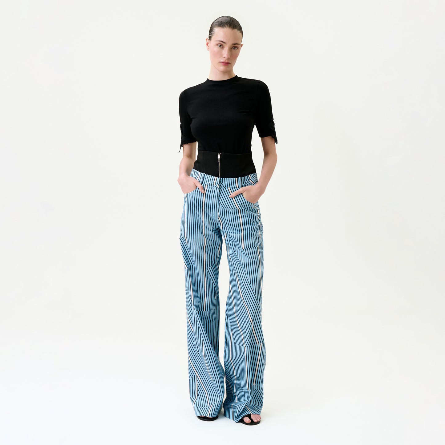 A woman stands against a white background wearing the Ruelle Pant—high-waisted, wide-leg blue and white striped 100% cotton pants—with her hands in her pockets and a fitted black top.