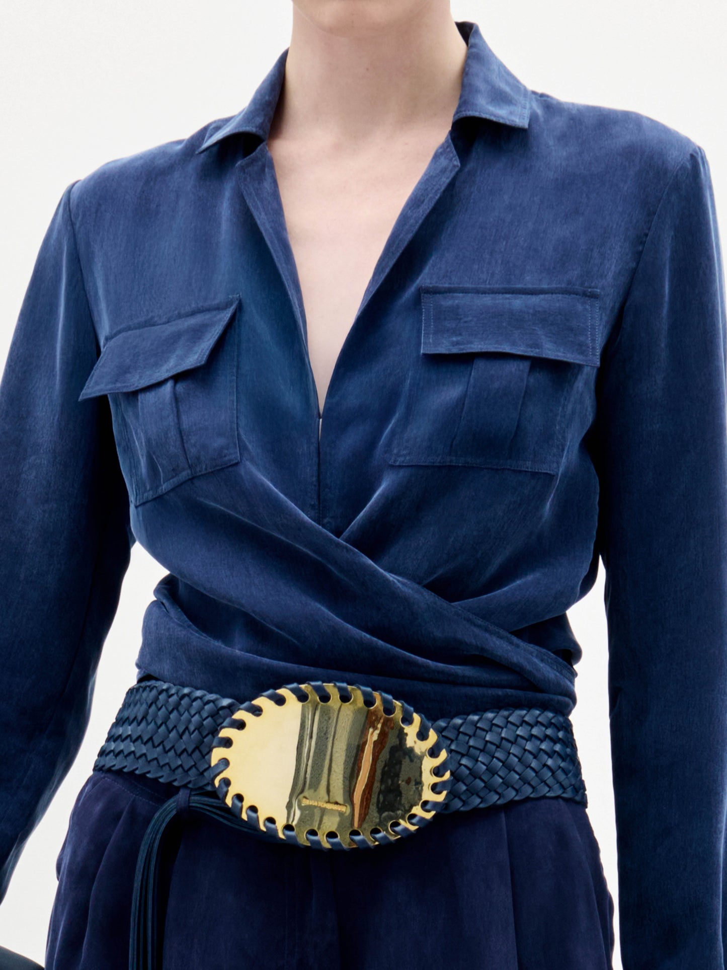 A woman wearing the Caserta Blouse in deep blue, paired with trousers, a gold-buckled belt, and a matching navy clutch, stands against a plain light background.