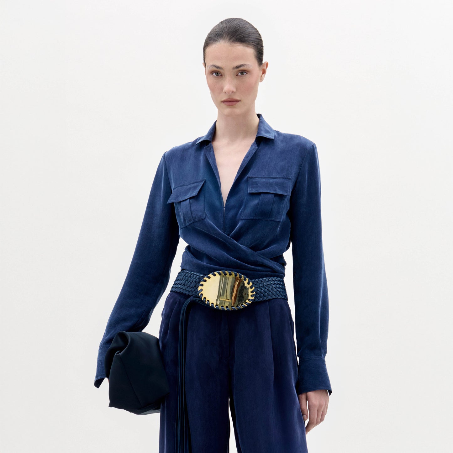 A woman wearing the Caserta Blouse in deep blue, paired with trousers, a gold-buckled belt, and a matching navy clutch, stands against a plain light background.