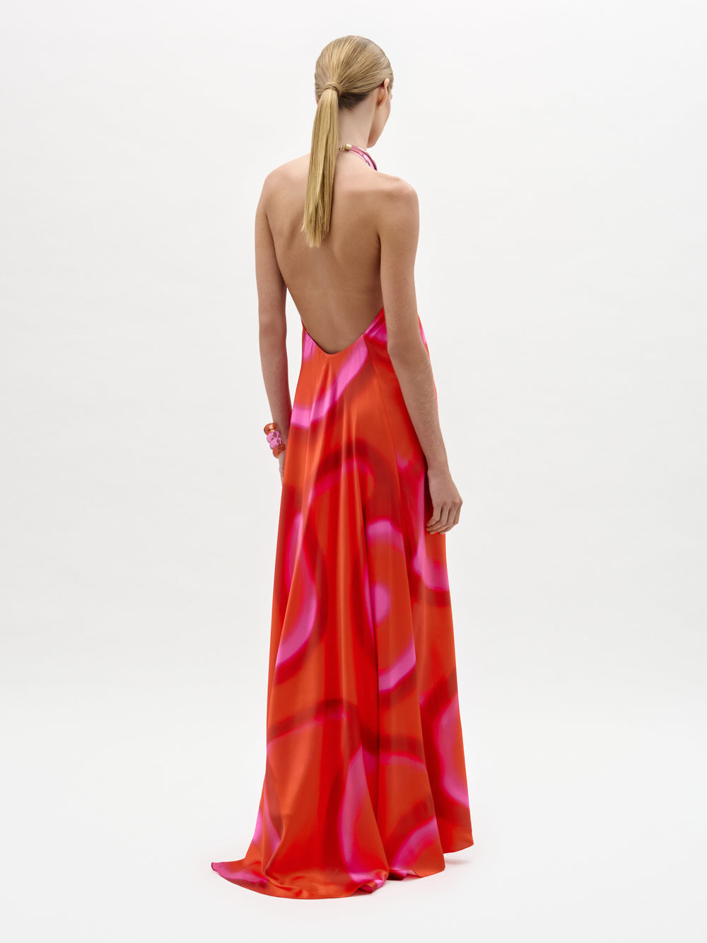 A person with blonde hair in a low ponytail wears the Jesse Dress—a backless, floor-length maxi with a halter neckline and a pink and red abstract pattern, made from luxurious Italian fabric—standing against a white background.