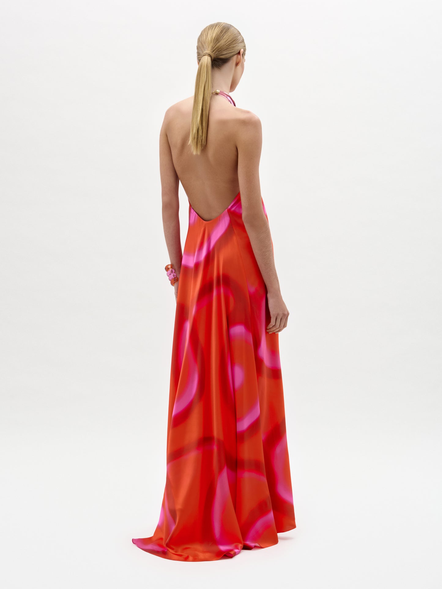 A person with blonde hair in a low ponytail wears the Jesse Dress—a backless, floor-length maxi with a halter neckline and a pink and red abstract pattern, made from luxurious Italian fabric—standing against a white background.