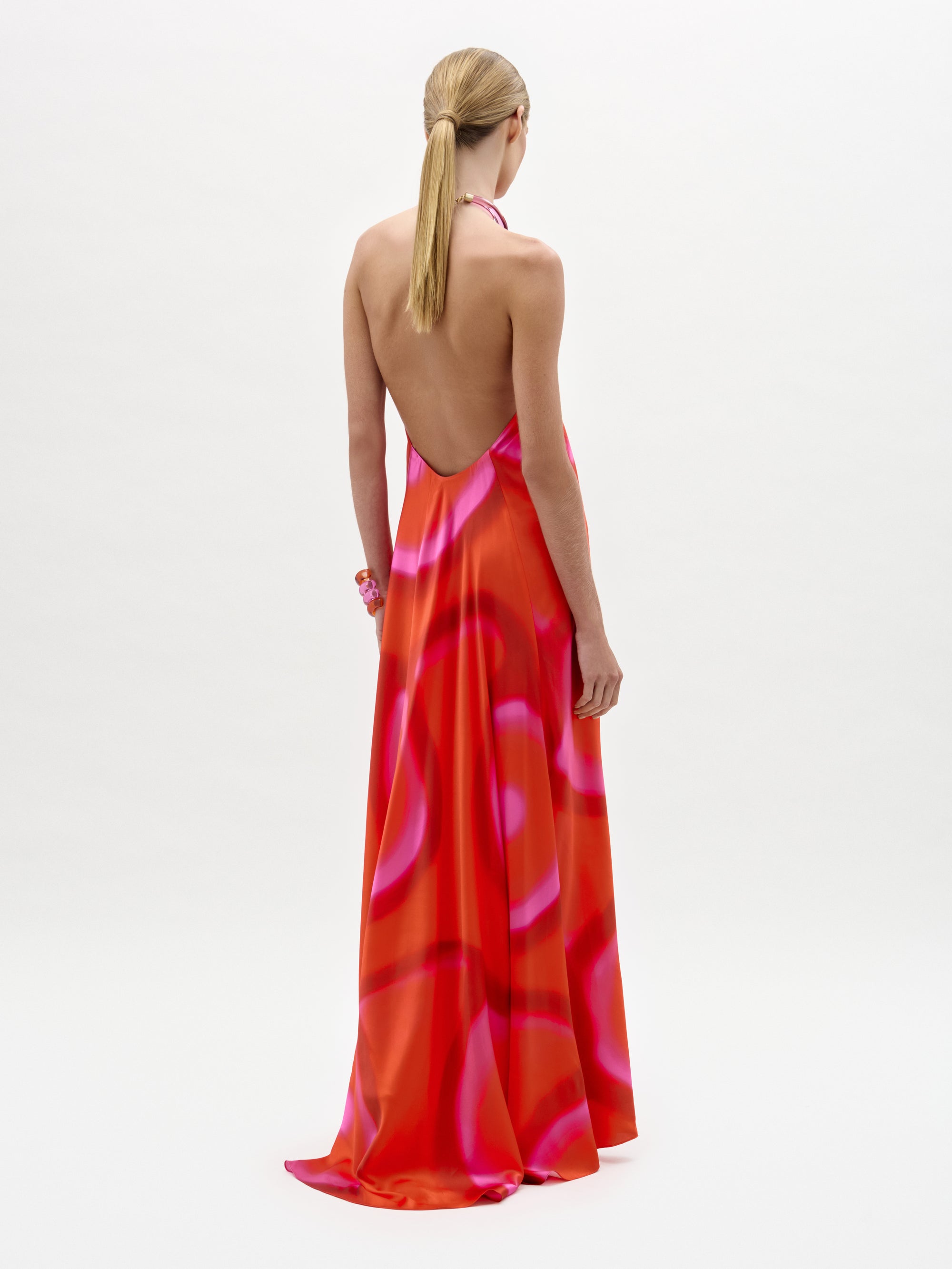 A person with blonde hair in a low ponytail wears the Jesse Dress—a backless, floor-length maxi with a halter neckline and a pink and red abstract pattern, made from luxurious Italian fabric—standing against a white background.