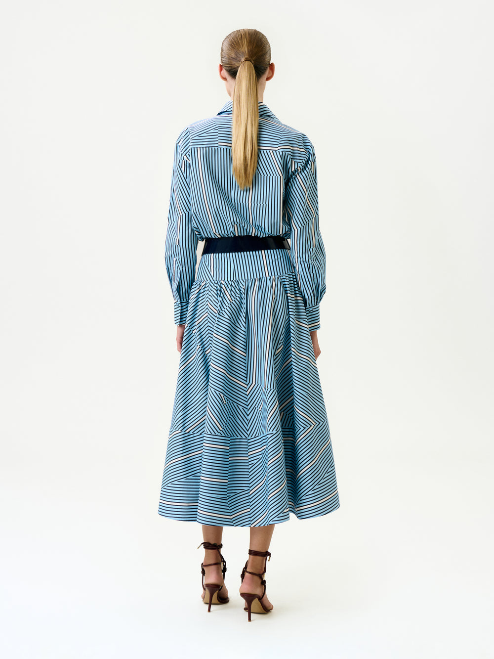 A person with long blond hair in a low ponytail wears the Milva Skirt, styled with a black belt and brown high-heeled sandals, facing away against a plain white background.
