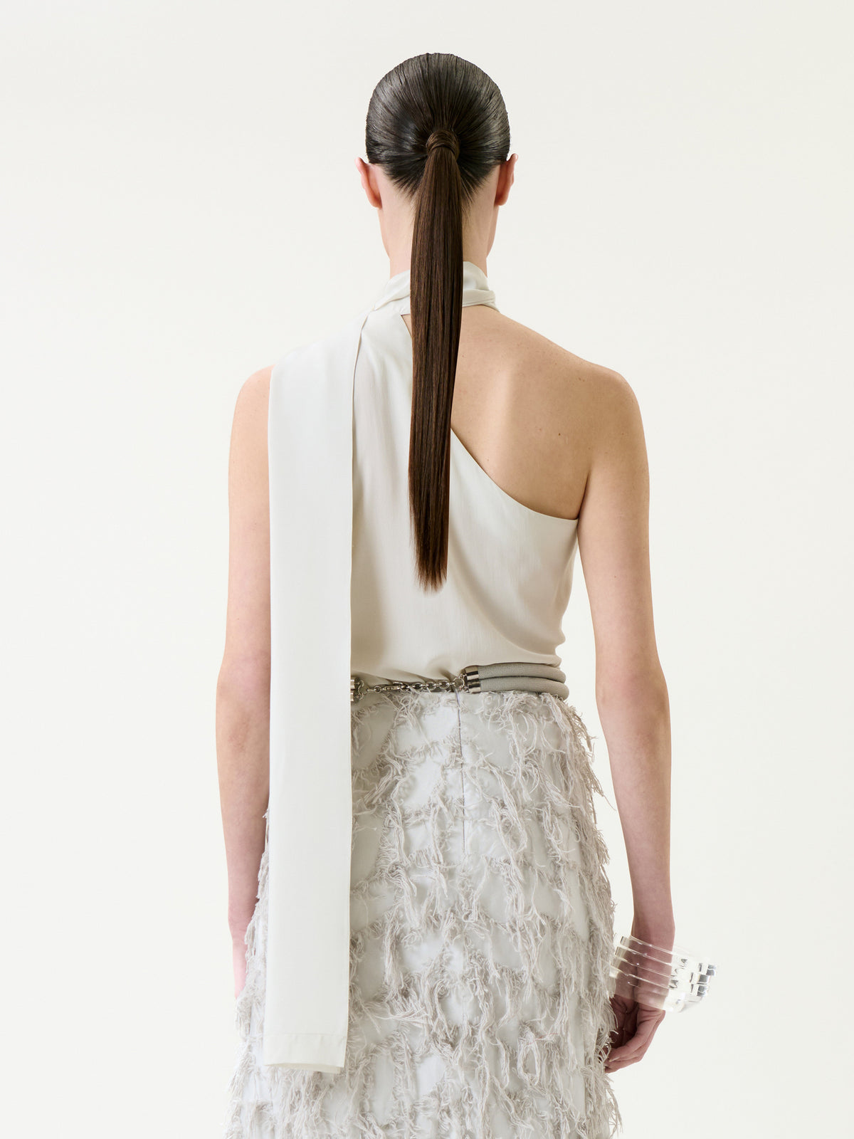A person with long, straight brown hair in a low ponytail is shown from behind against a plain light background, wearing the Onora Blouse—an elegant one-shoulder white top paired with a textured white skirt.