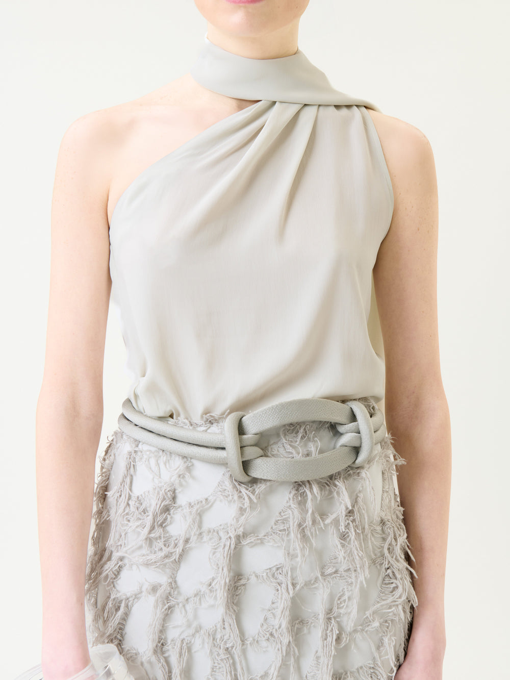 Wearing the Onora Blouse in light gray, accented by a matching wide belt and paired with a textured, fringed skirt, the person embodies a soft and elegant look. Their face is partially out of frame.
