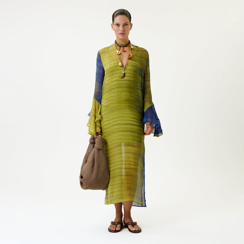 A person wearing the Aries Dress, a sheer green and blue long-sleeve dress, along with brown sandals, a necklace, and holding a large brown textured bag, stands against a plain white background.