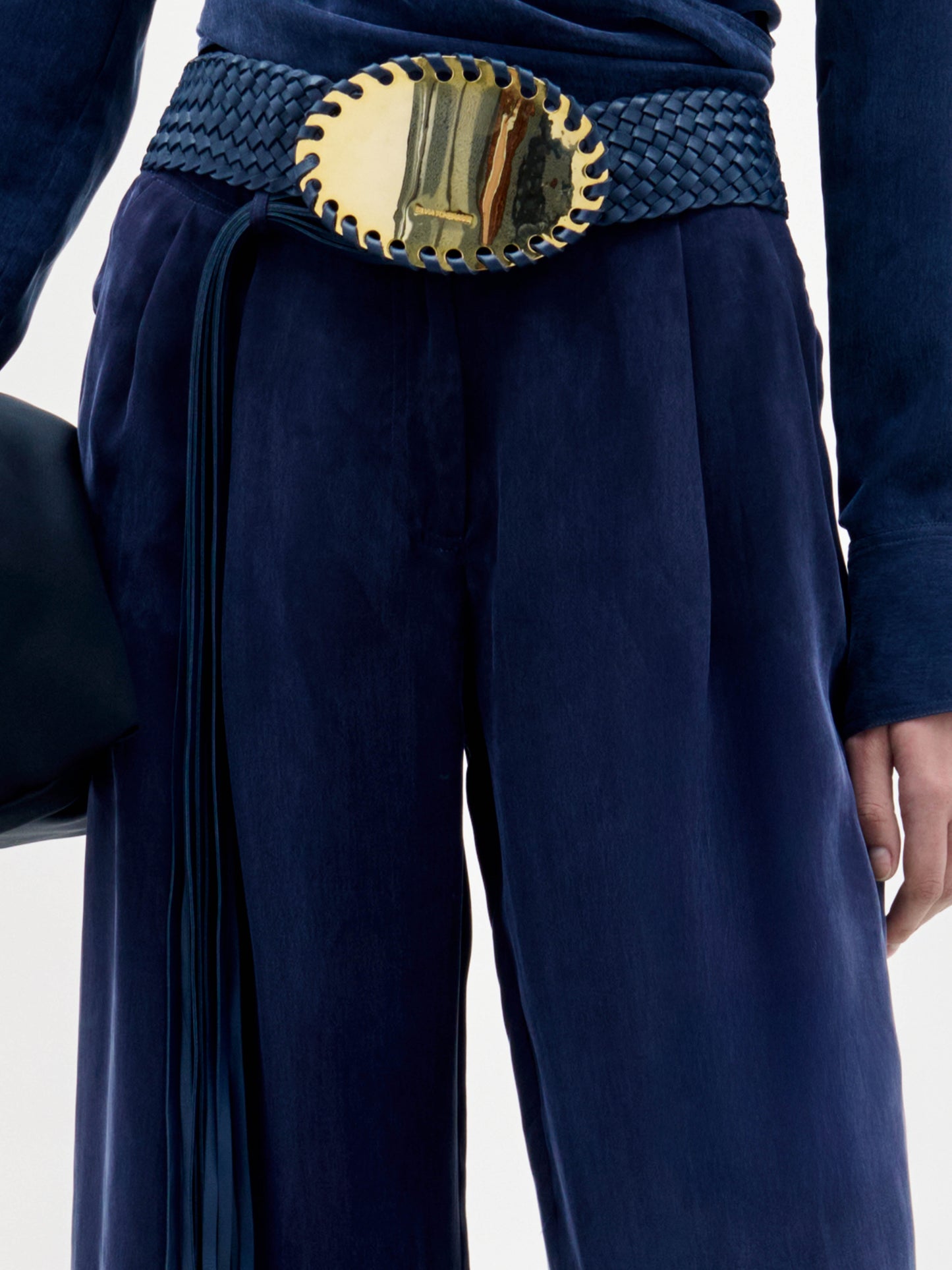 A woman stands against a plain background wearing the Opal Pant, paired with a deep blue long-sleeve blouse, a large gold-detailed belt, sandals, and holding a small clutch.