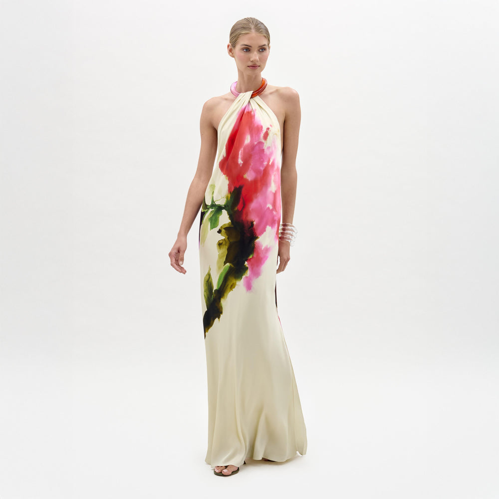 A woman stands against a plain background wearing the Jesse Dress, a sleeveless cream maxi with a colorful abstract floral print and halter neckline, paired with open-toe heels.