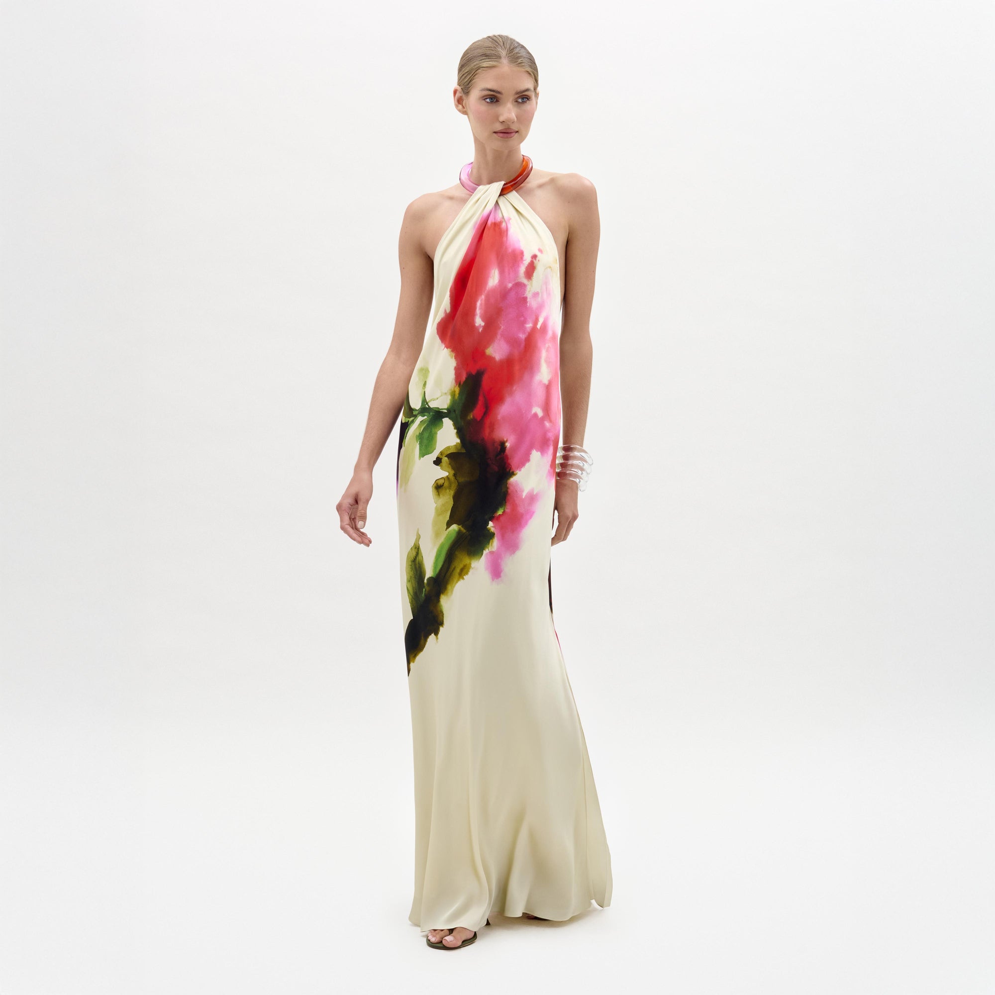 A woman stands against a plain background wearing the Jesse Dress, a sleeveless cream maxi with a colorful abstract floral print and halter neckline, paired with open-toe heels.