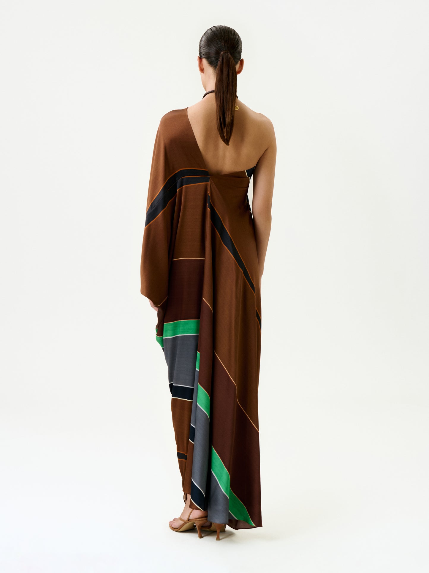 A woman with dark hair in a low ponytail stands facing away, wearing the Inu Tunic—a silk brown one-shoulder maxi dress with black, green, and gray stripes—paired with tan sandals, against a plain white background.