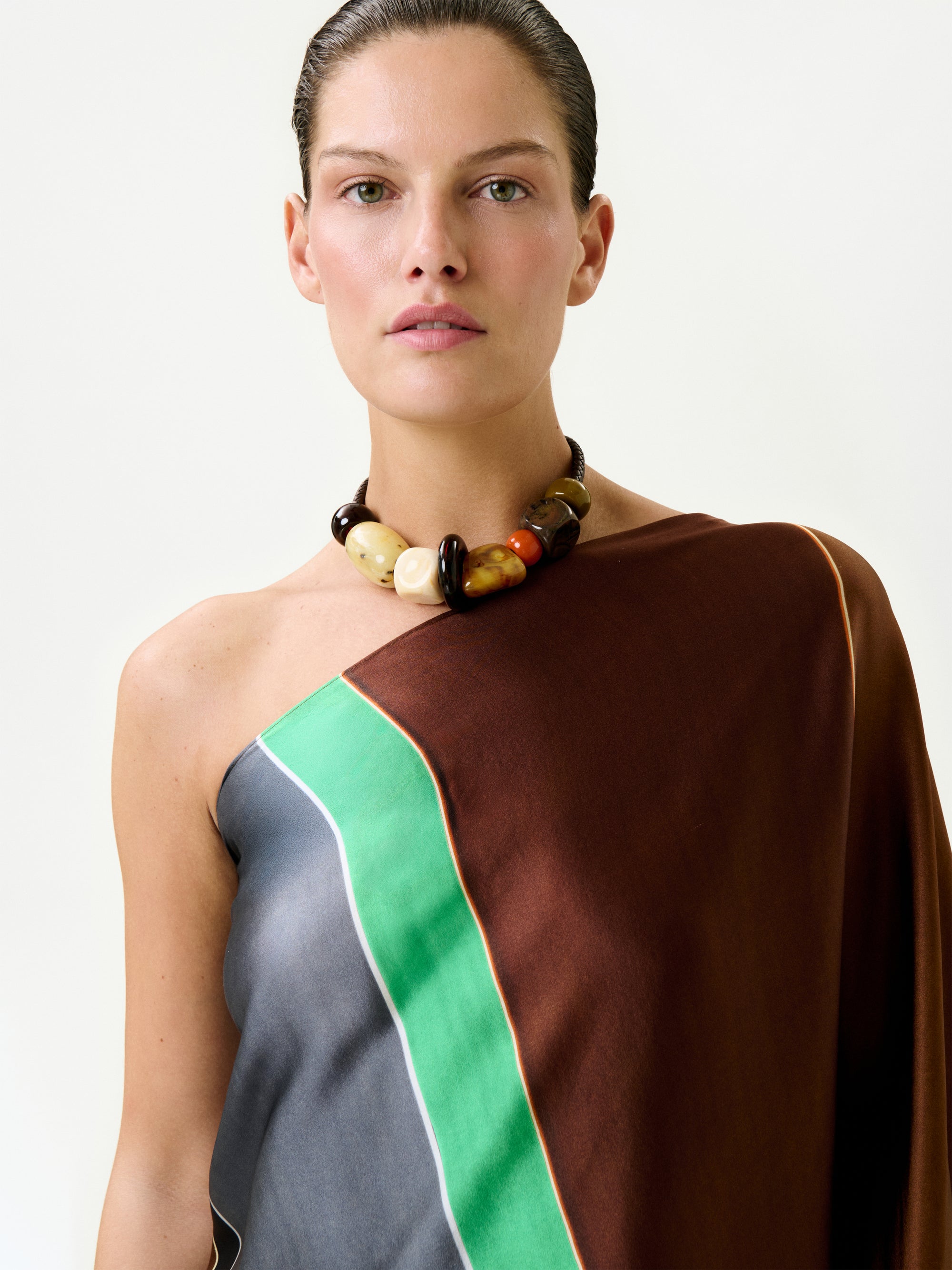 A woman with slicked-back hair wears the Inu Tunic—a one-shoulder silk maxi dress in brown, green, and gray hues—and a chunky beaded necklace, standing against a plain light background.