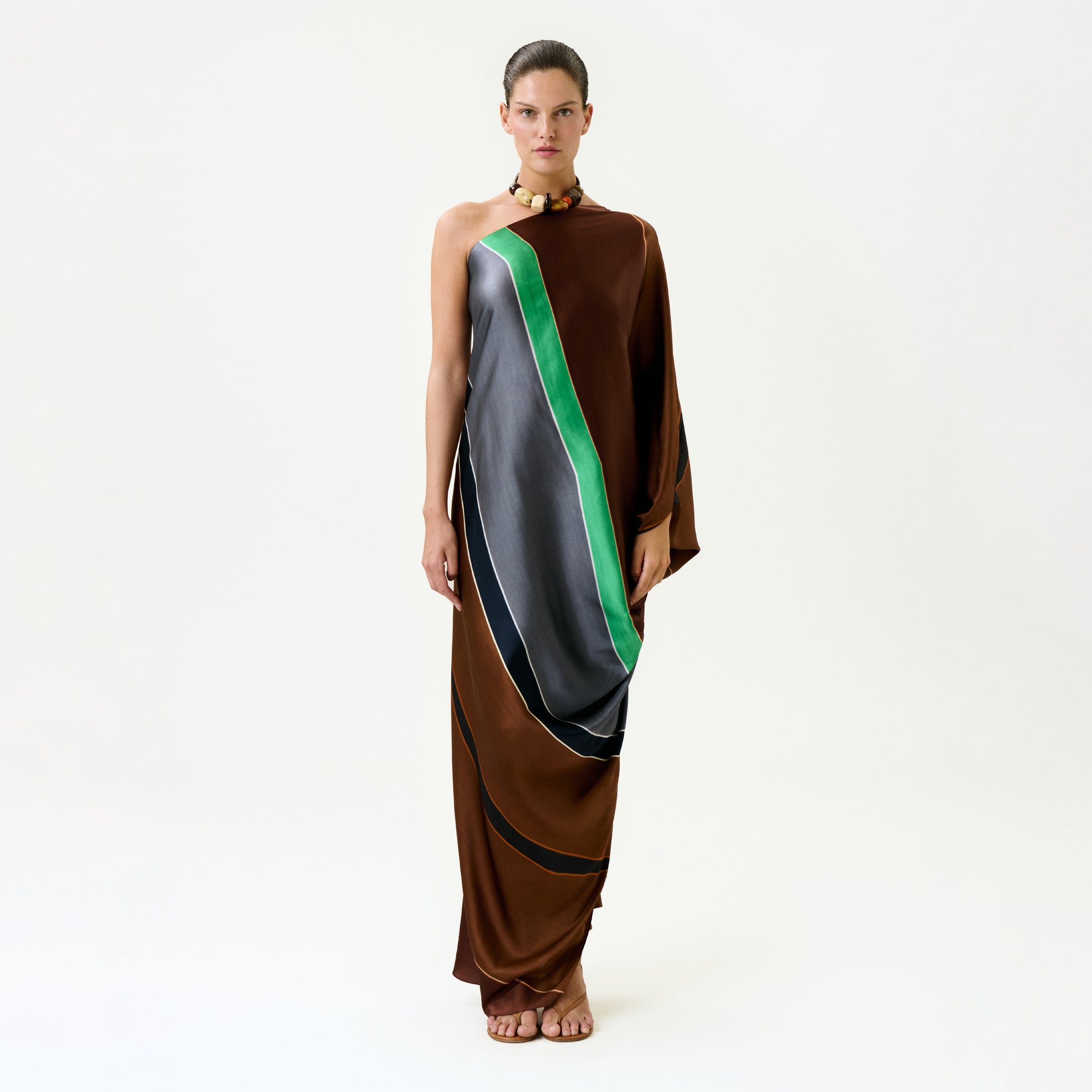 A woman stands against a plain white background wearing the Inu Tunic, a one-shoulder maxi dress with green, gray, and black diagonal stripes in luxurious Italian fabric. She is barefoot, hair pulled back, and wears a necklace.