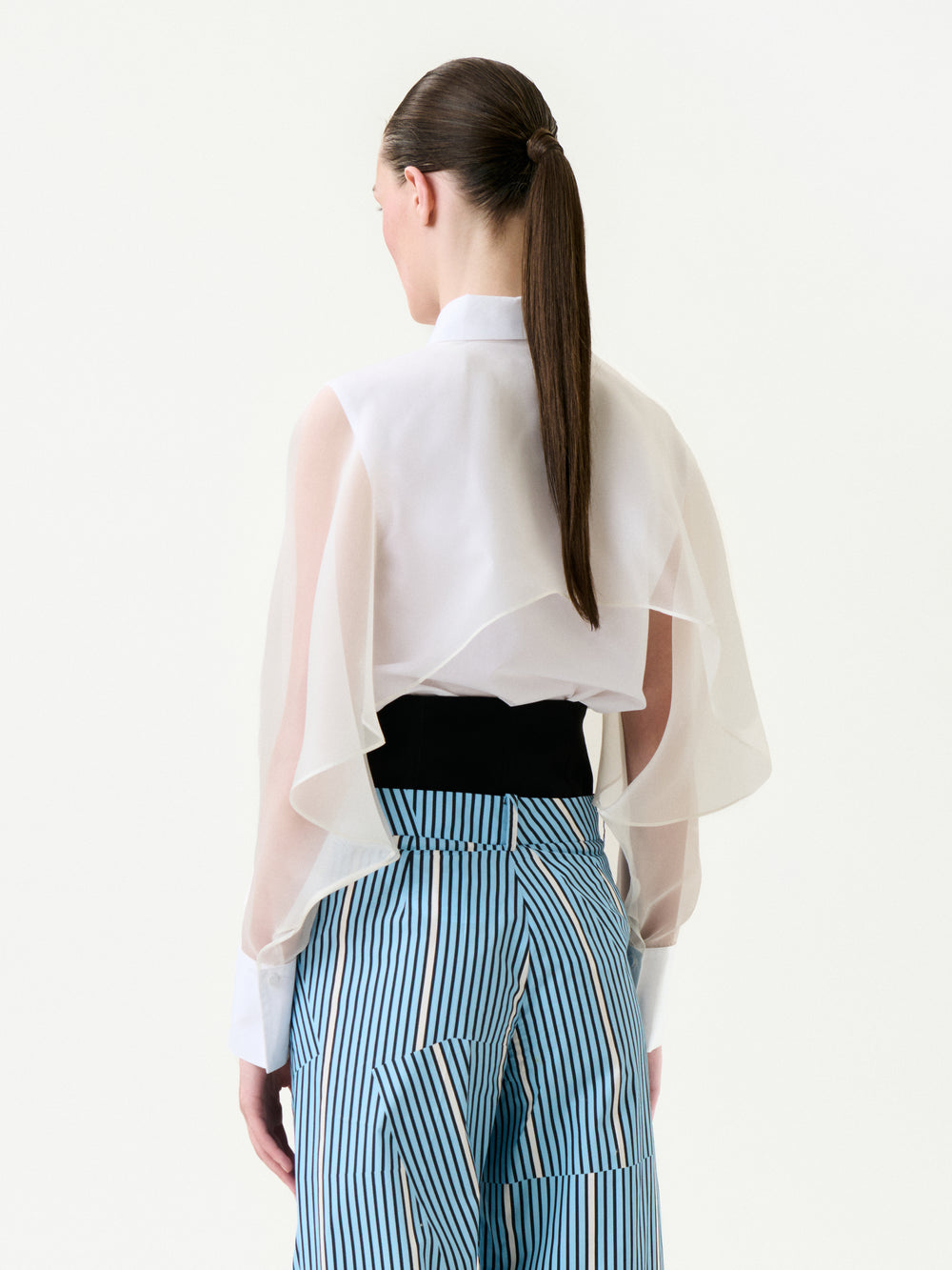 A person faces away, wearing the Nira Shirt—a sheer white blouse with flowing sleeves—tucked into high-waisted blue and white striped cotton pants. Their long brown hair is styled in a sleek ponytail.