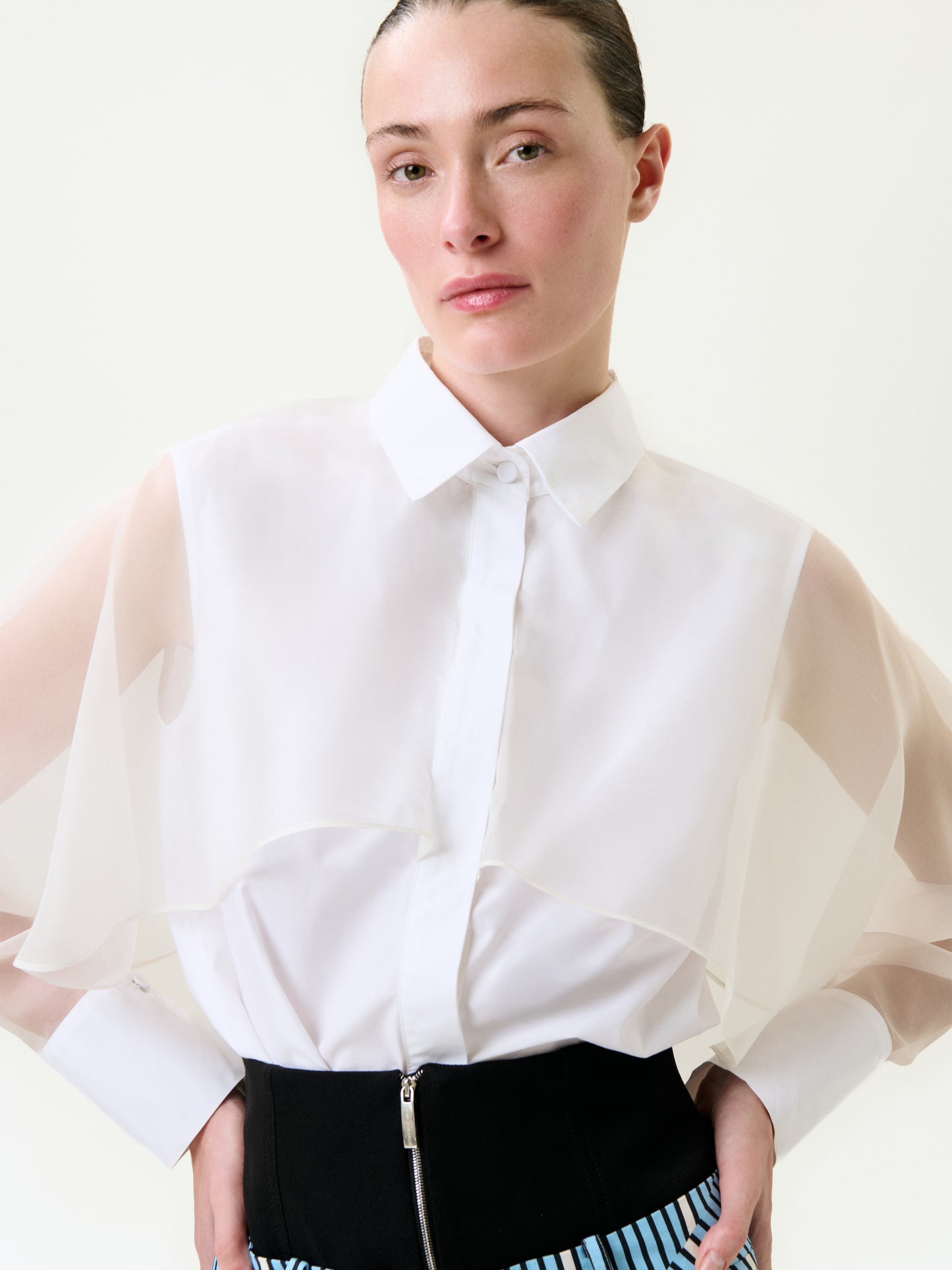 A woman with slicked-back hair wears the Nira Shirt—a sheer, layered-sleeve blouse made from 100% cotton Portuguese fabric—and a high-waisted black skirt with blue and white stripes, standing before a plain light background.
