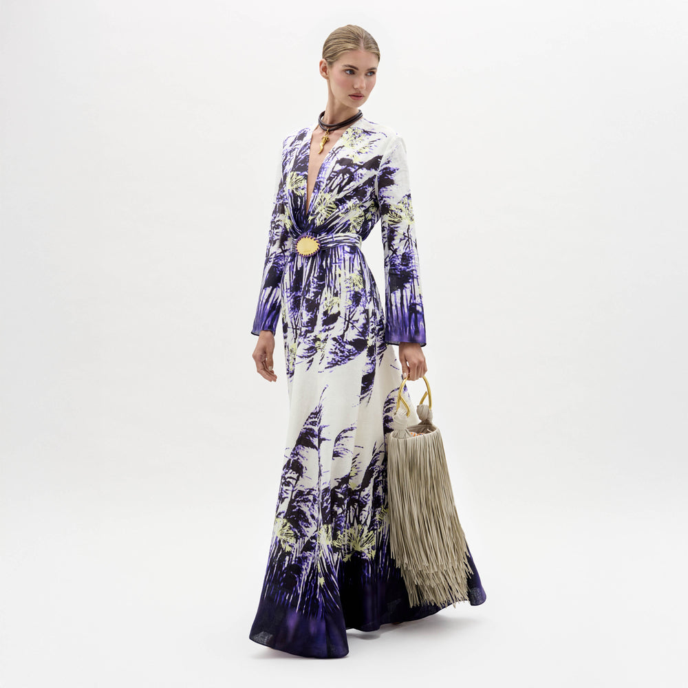 A woman in the Kady Dress, a long patterned purple and white gown, stands holding a large beige fringed handbag against a plain white background.