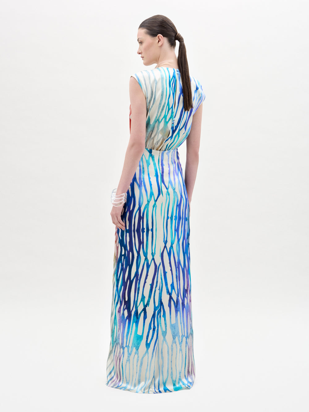 A woman stands against a plain background wearing the Samaira Dress, a sleeveless maxi with a ruched waist and a vibrant red, blue, yellow, and white abstract pattern.