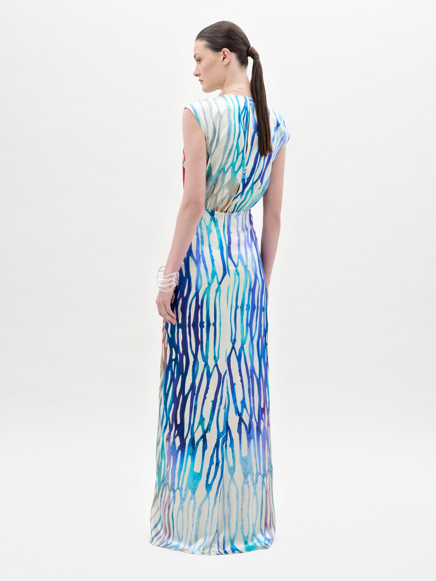 A woman stands against a plain background wearing the Samaira Dress, a sleeveless maxi with a ruched waist and a vibrant red, blue, yellow, and white abstract pattern.