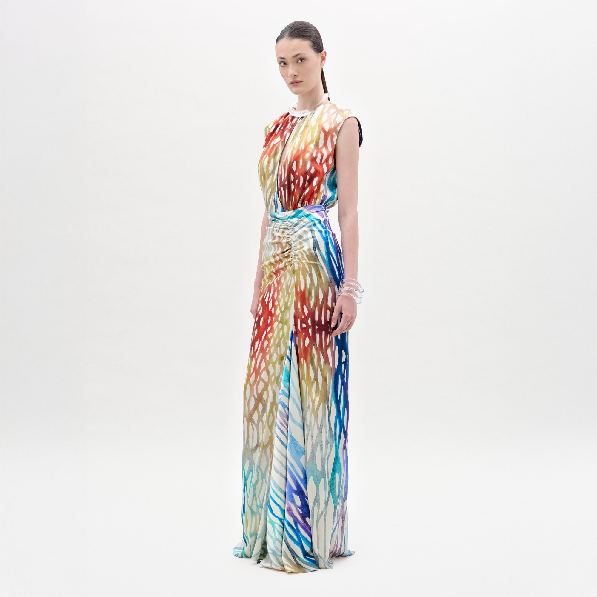 A woman stands against a plain background wearing the Samaira Dress, a sleeveless maxi with a ruched waist and a vibrant red, blue, yellow, and white abstract pattern.