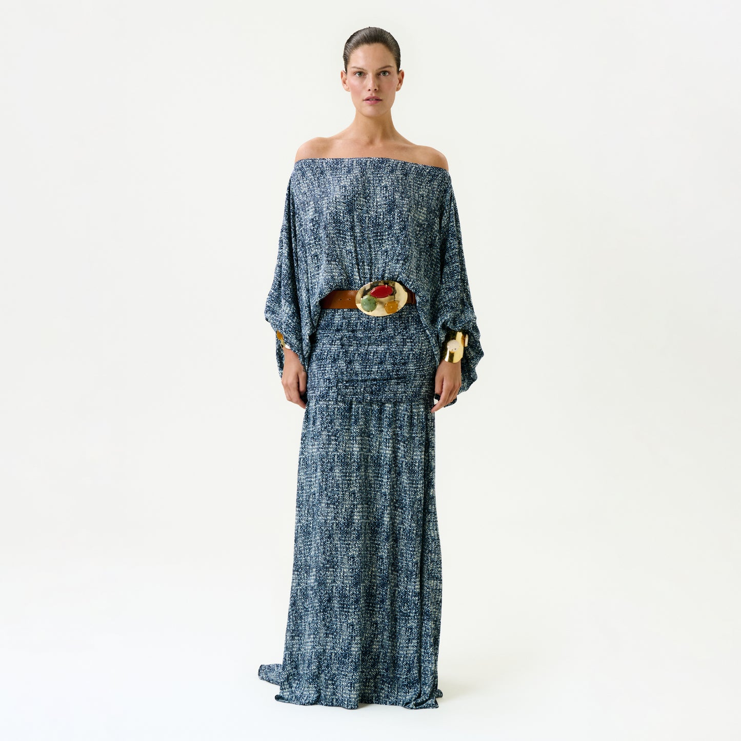 A woman faces forward wearing the Nelly Dress, a blue and white off-the-shoulder maxi made from Italian fabric. Her hair is pulled back and she accessorizes with a large brown belt with a colorful, decorative buckle.