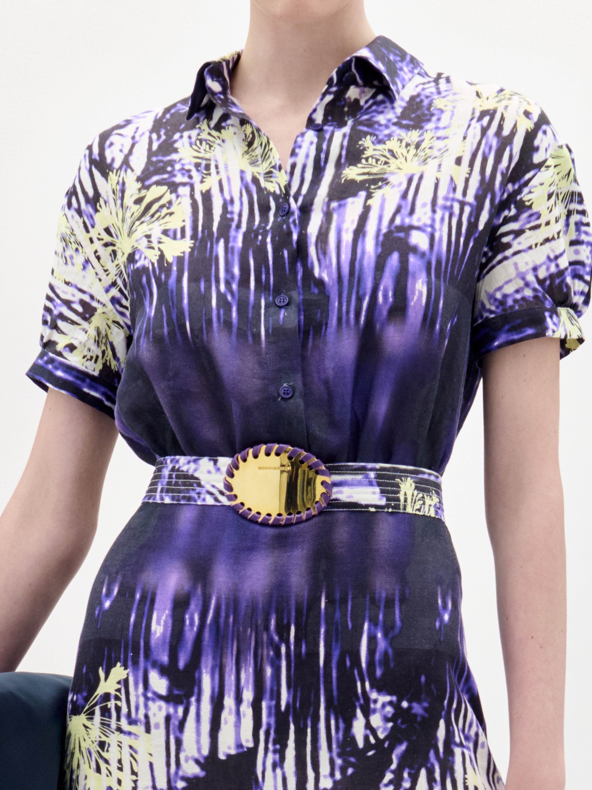 A woman wears the Lee Dress, a short-sleeve purple and white patterned dress with a belt, paired with a black clutch and black sandals, standing against a plain white background.