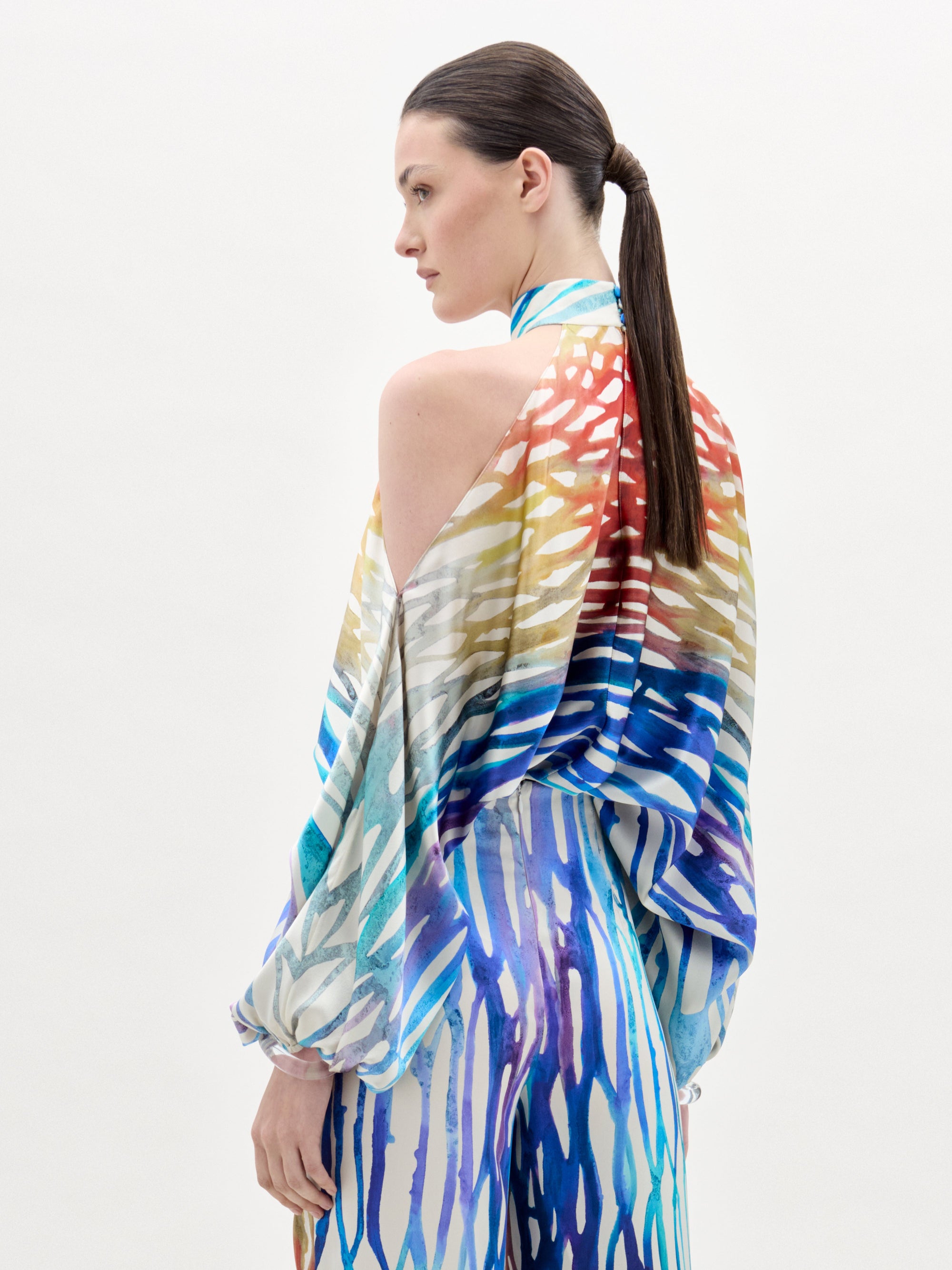 A woman models the Safira Blouse—an off-shoulder top featuring a colorful, abstract print in luxe Italian fabric—paired with matching wide-leg pants, set against a plain white background.