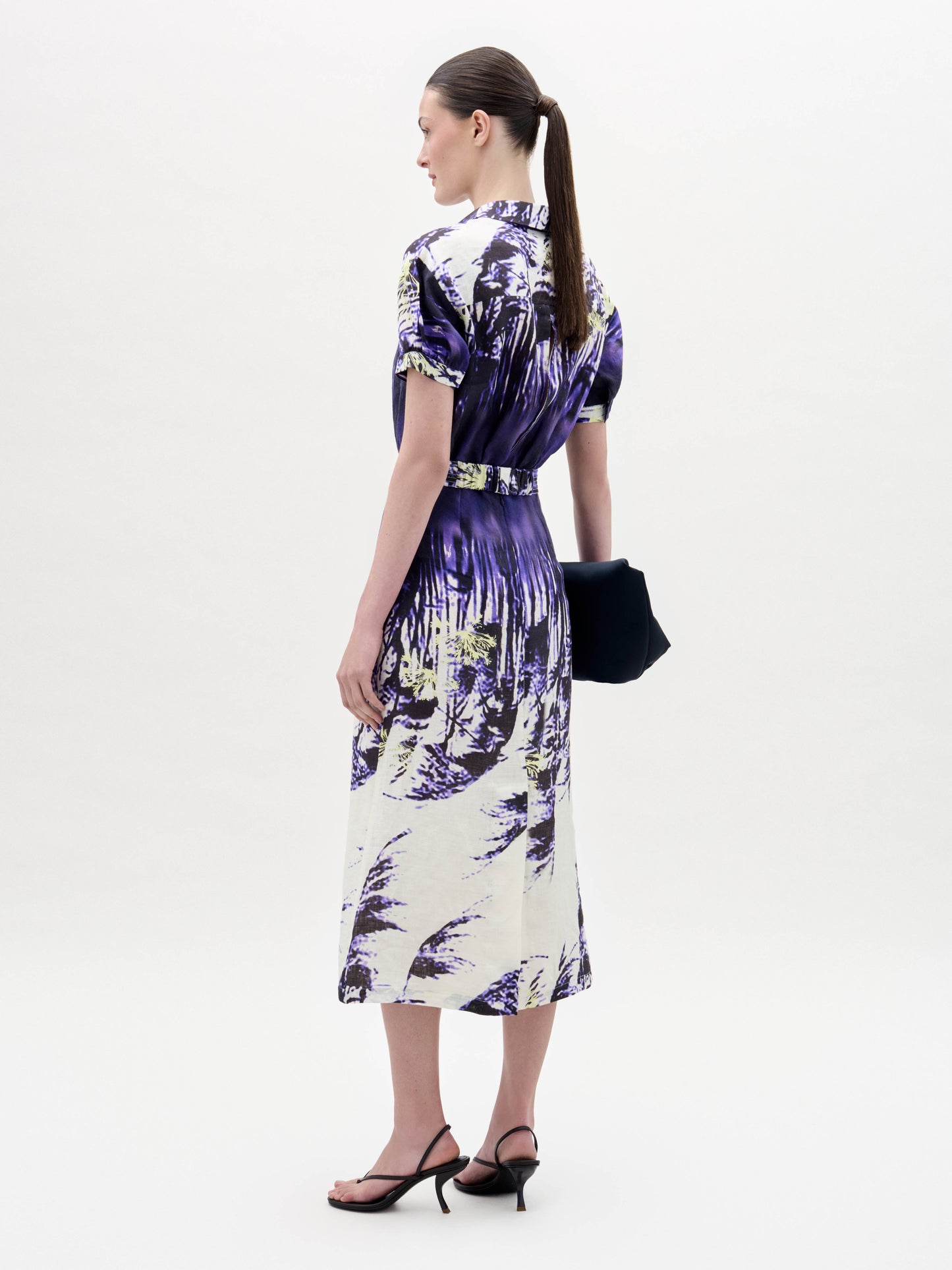 A woman wears the Lee Dress, a short-sleeve purple and white patterned dress with a belt, paired with a black clutch and black sandals, standing against a plain white background.