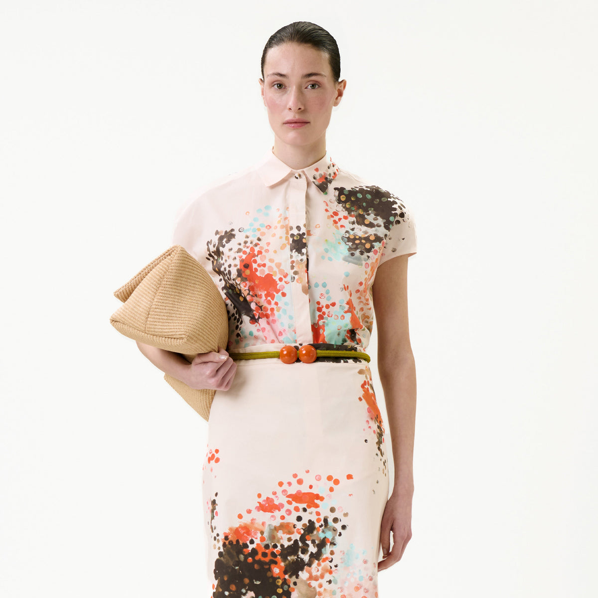 A woman wears the Maribel Shirt, a short-sleeve top featuring an orange, brown, and aqua spot pattern. She holds a tan textured clutch, has her hair pulled back, and accessorizes with a belt adorned with two orange beads.